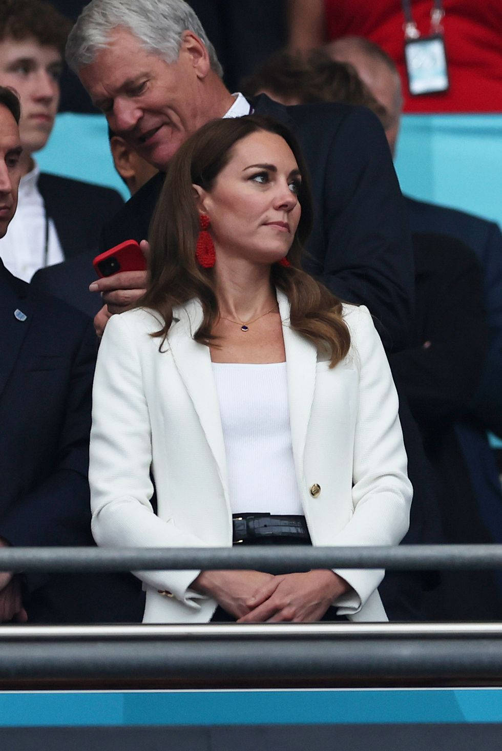 Kate Middleton 桌布 壁紙 — 凱特·米德爾頓身穿白色西裝外套和紅色耳環，在體育場座位上專注地觀看。