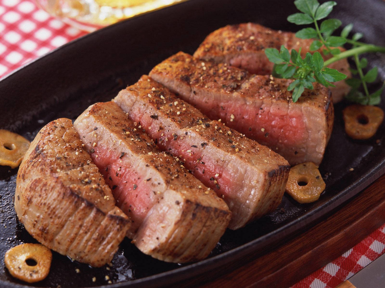 meat 桌布 壁紙 — 切片中等熟度牛排，帶焦脆外皮和新鮮香草，放在黑色盤子上，配橄欖和格子桌布。