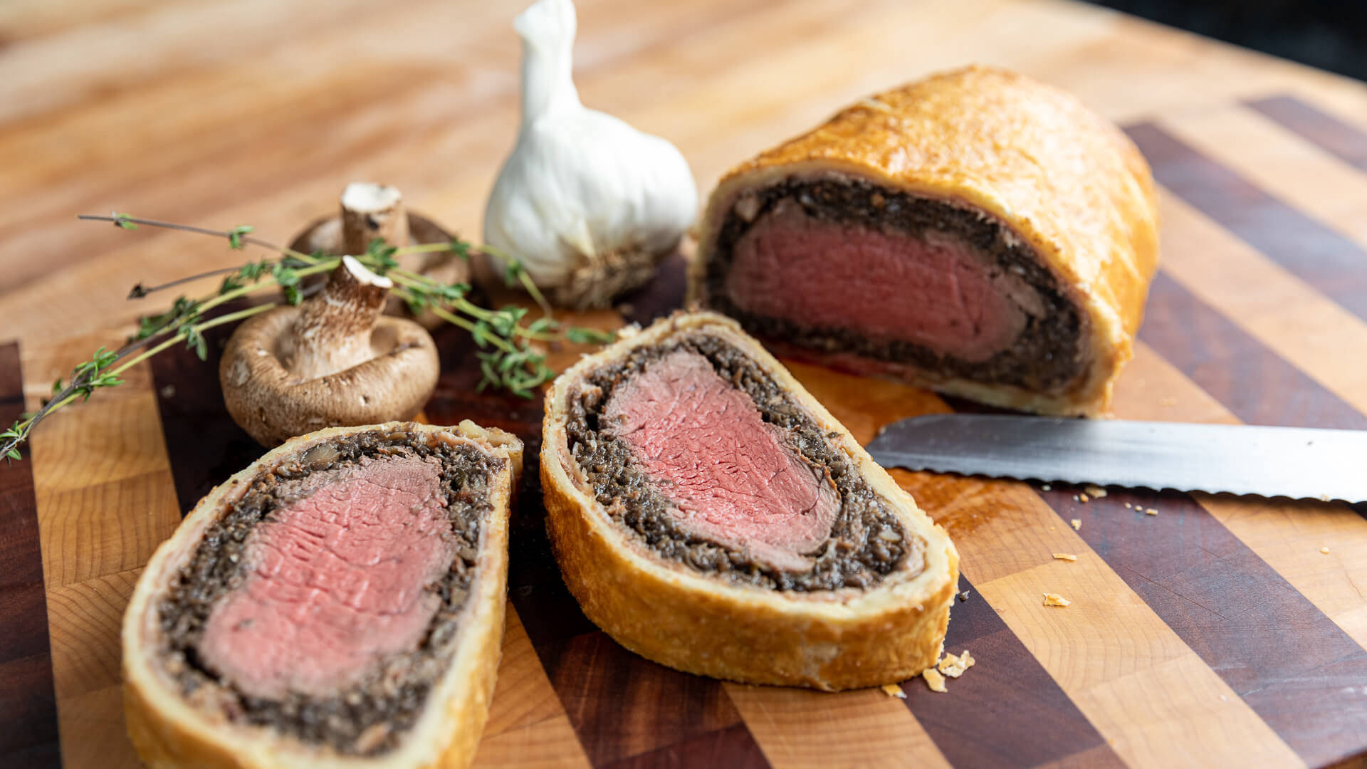 meat 桌布 壁紙 — 切片牛肉威靈頓配金黃酥皮和蘑菇醬，放在條紋木砧板上，旁邊有蒜頭百里香和一把刀。
