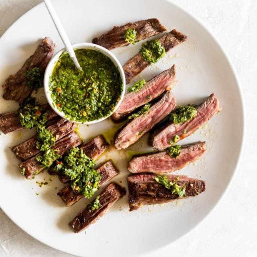 meat 桌布 壁紙 — 切片的中等熟度牛排排列在白色盤子上，搭配鮮綠色的 chimichurri 醬。