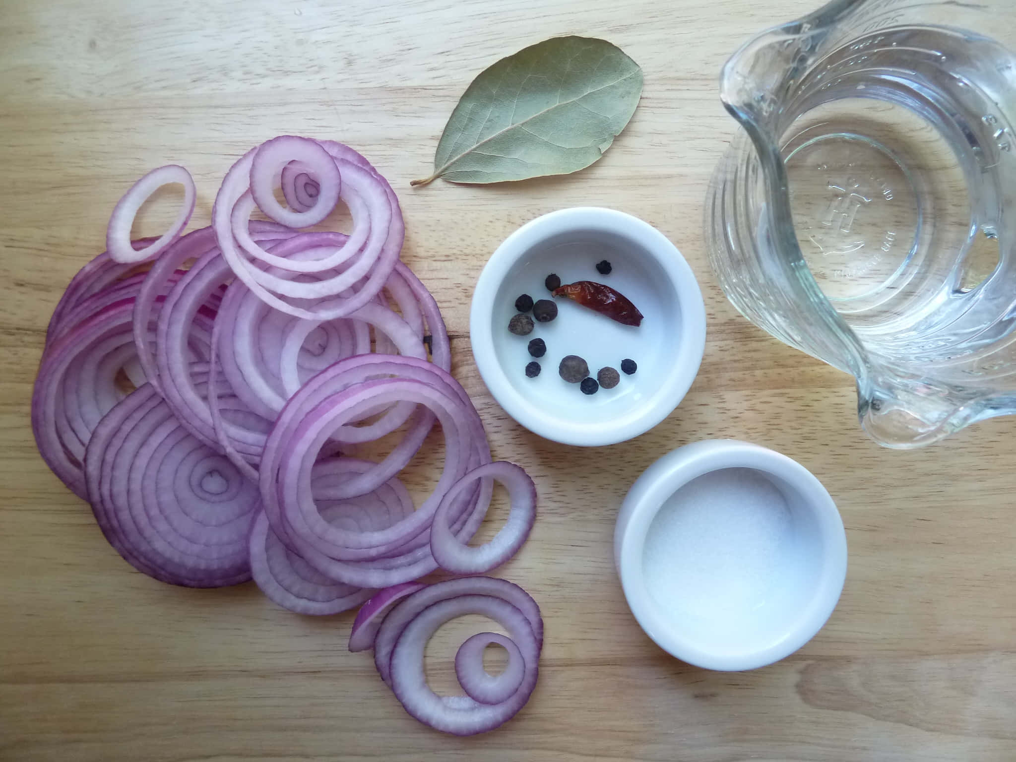Purple Onions 桌布 壁紙 — 切片紅洋蔥、胡椒粒、月桂葉和鹽擺放在木製砧板上，配有一個玻璃水壺。