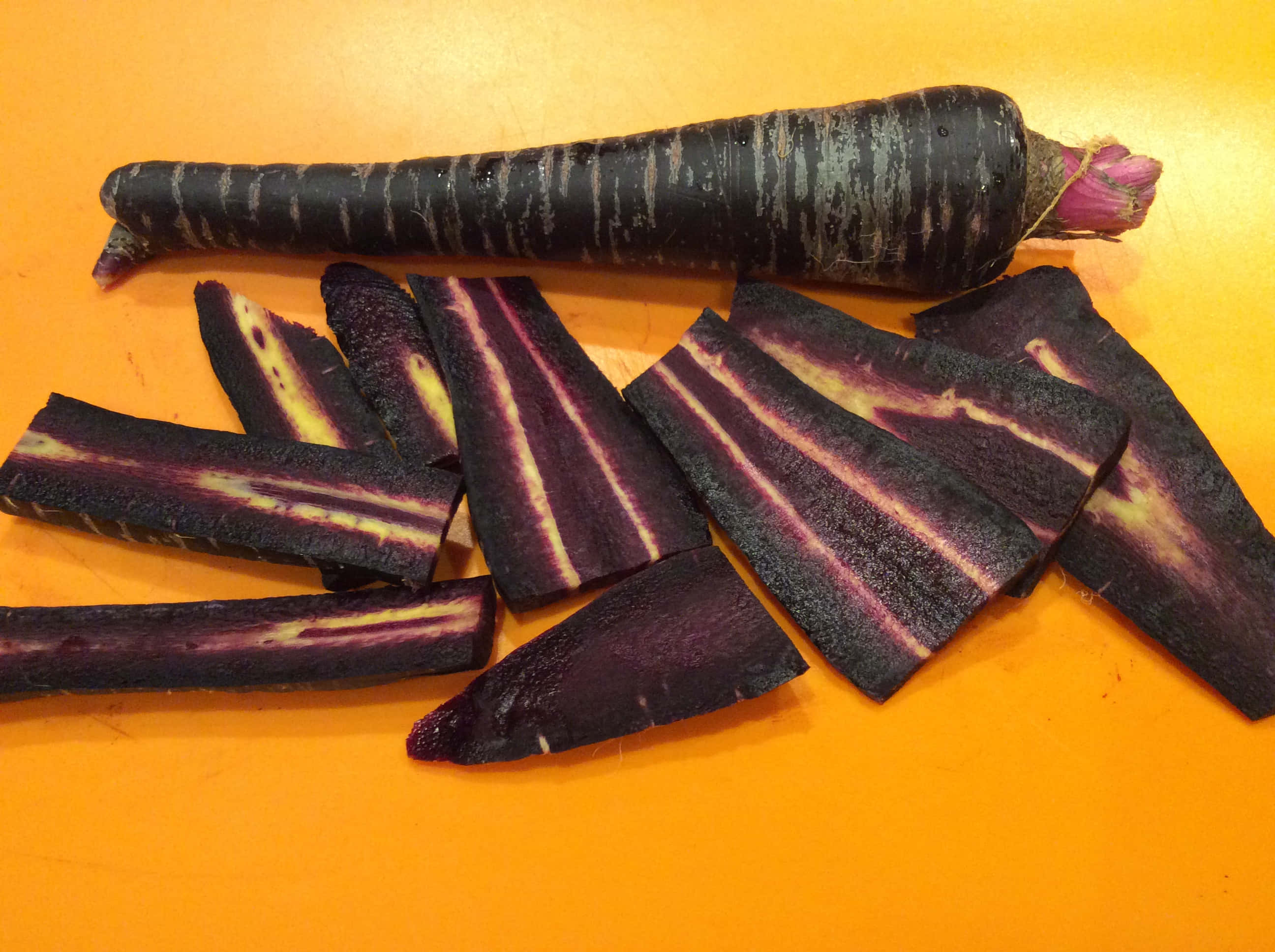 Purple Carrots 桌布 壁紙 — 切片黑蘿蔔帶有紫色和黃色條紋，放在亮橙色表面上，旁邊是完整的蘿蔔。