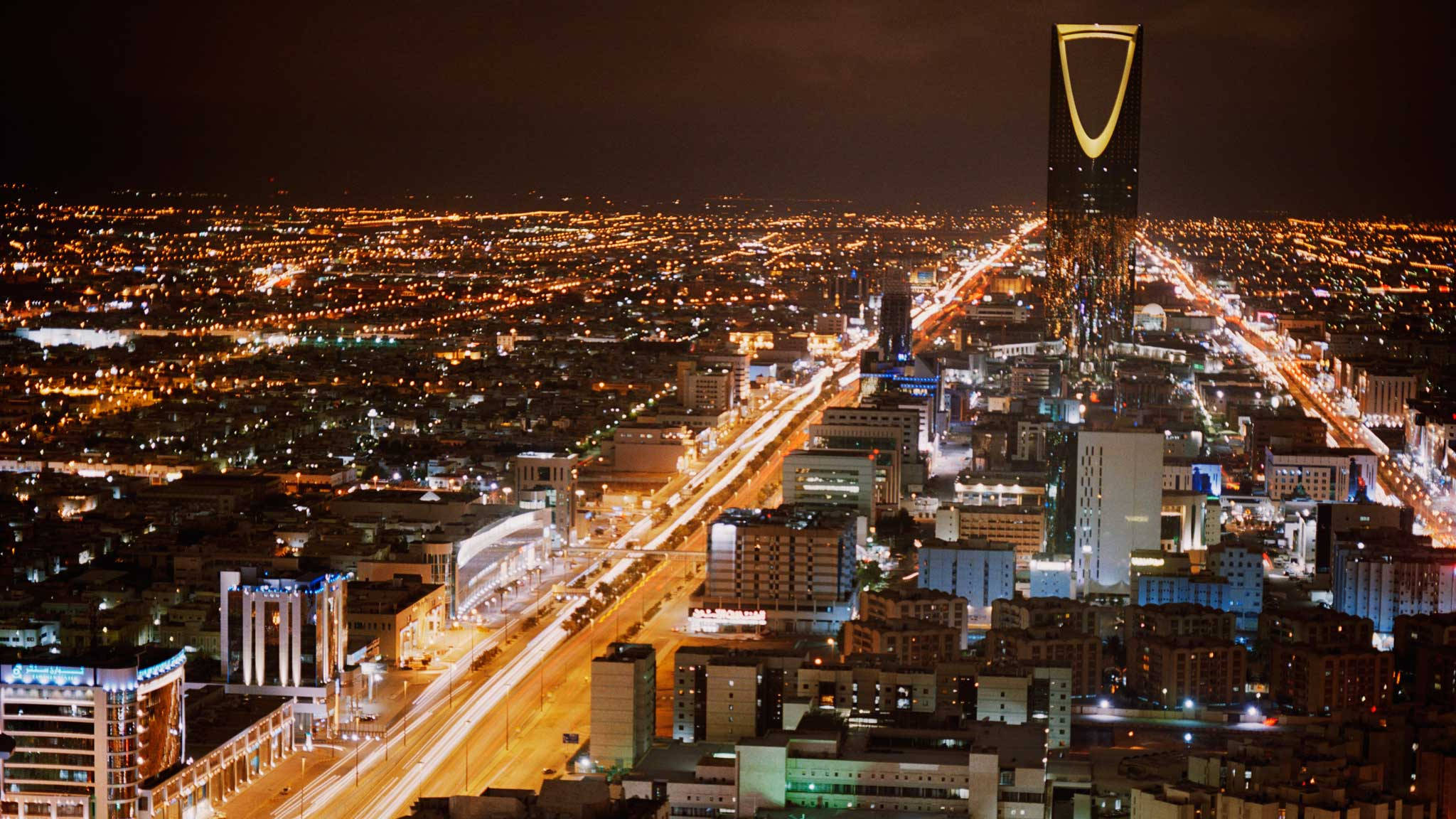 Riyadh 桌布 壁紙 — 利雅德城市景觀的空中夜景，金色路燈和發光的摩天大樓照亮，王國塔高聳入雲。