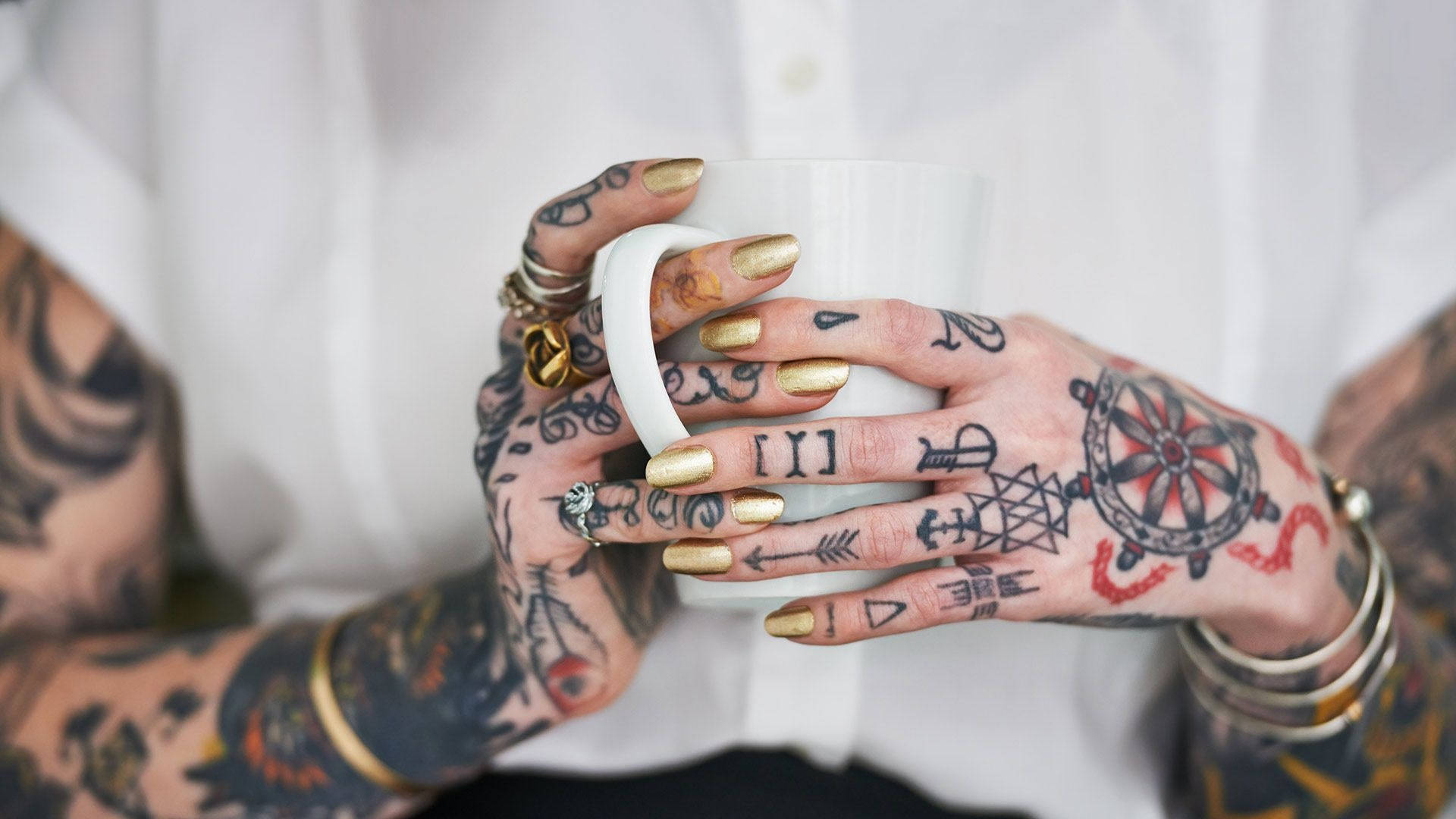 Tattoo 桌布 壁紙 — 刺青的手戴著金色指甲，捧著一個白色馬克杯，背景是柔軟的白色襯衫。