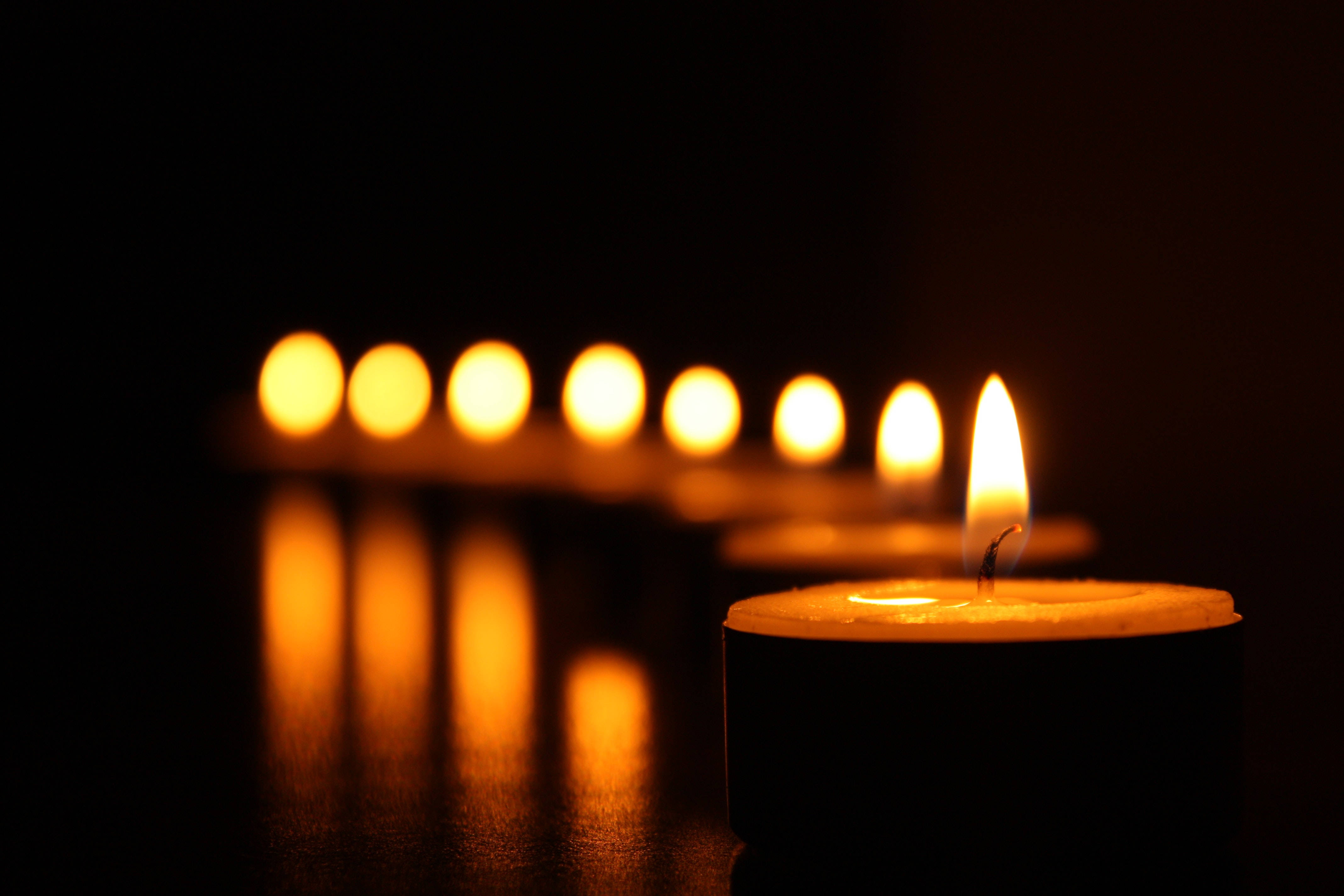 All Saints Day 桌布 壁紙 — 前景中一支點燃的茶燭散發溫暖光芒，背後一排模糊的蠟燭逐漸消失在黑暗中。