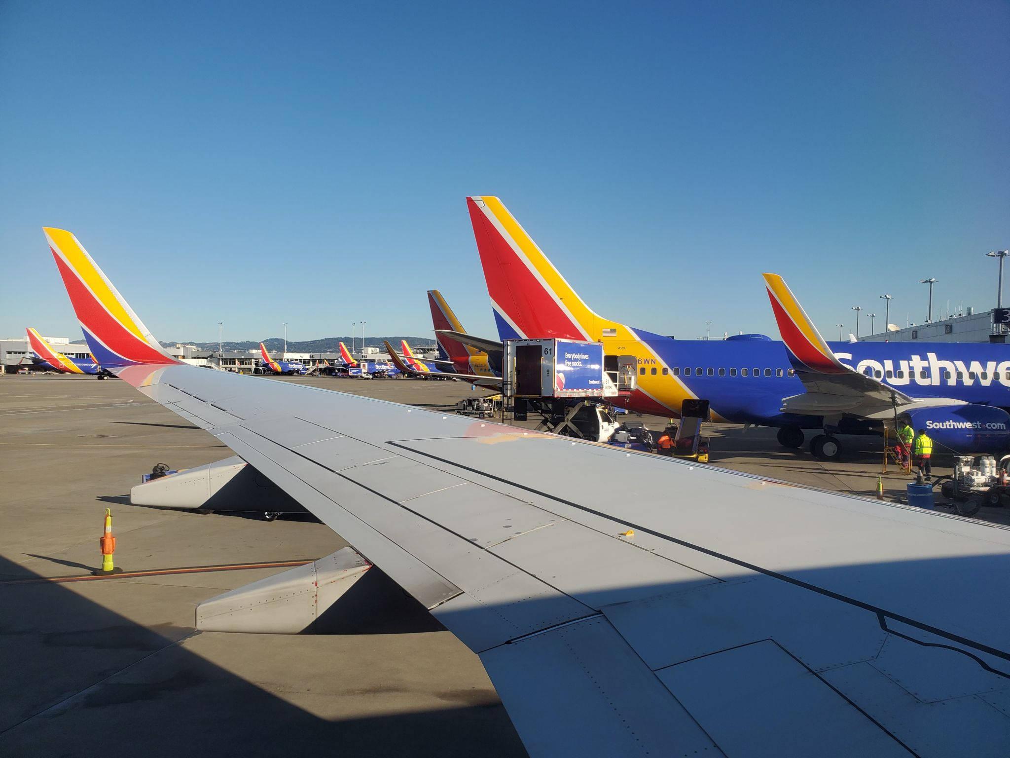 Southwest 桌布 壁紙 — 前景中的飛機機翼，西南航空飛機在晴朗藍天下停泊在機場停機坪閘口。