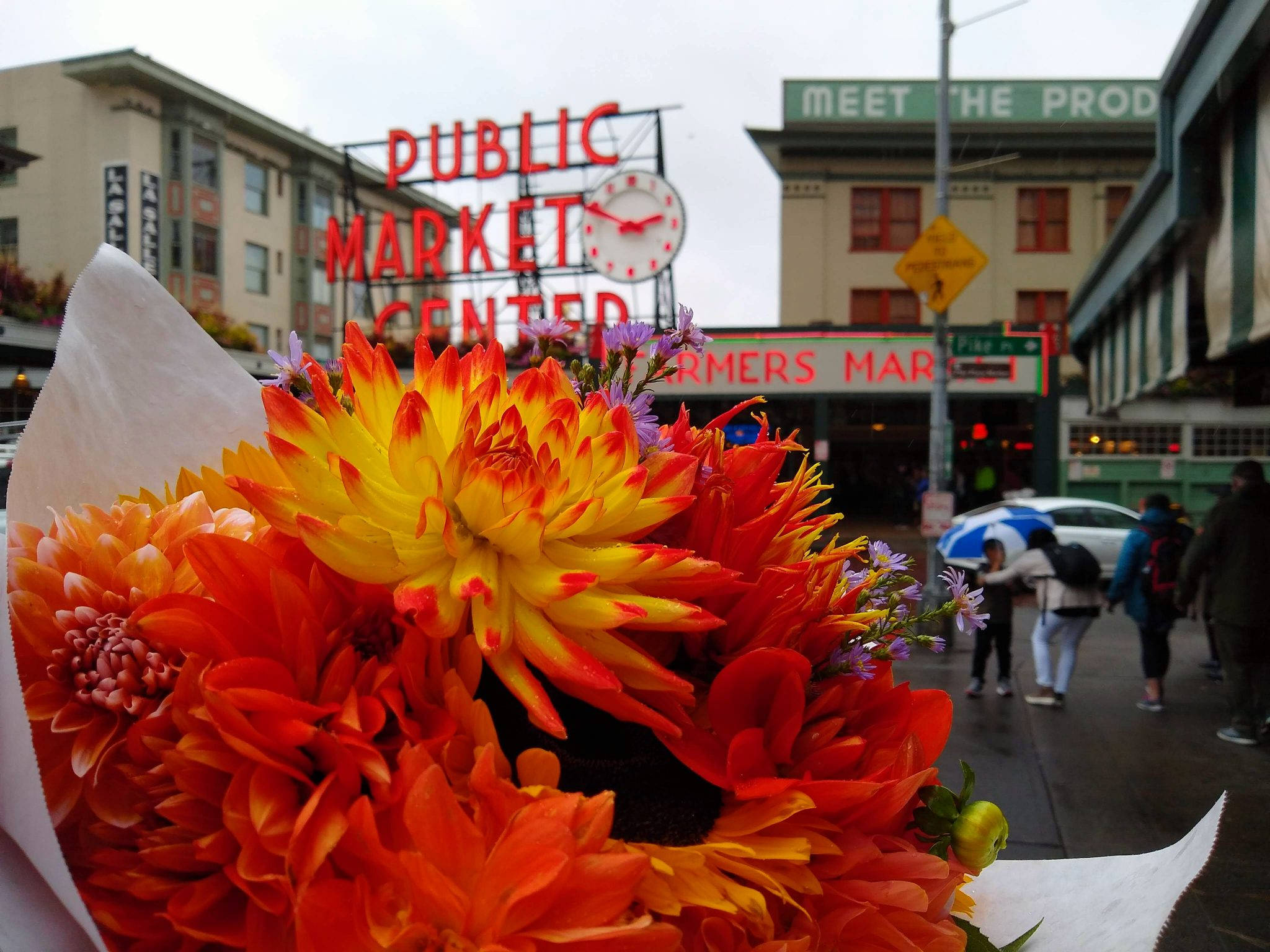Pike Place Market 桌布 壁紙 — 前景是鮮豔的橙色和黃色花朵，後面是西雅圖公共市場中心的標誌和雨中的街景。