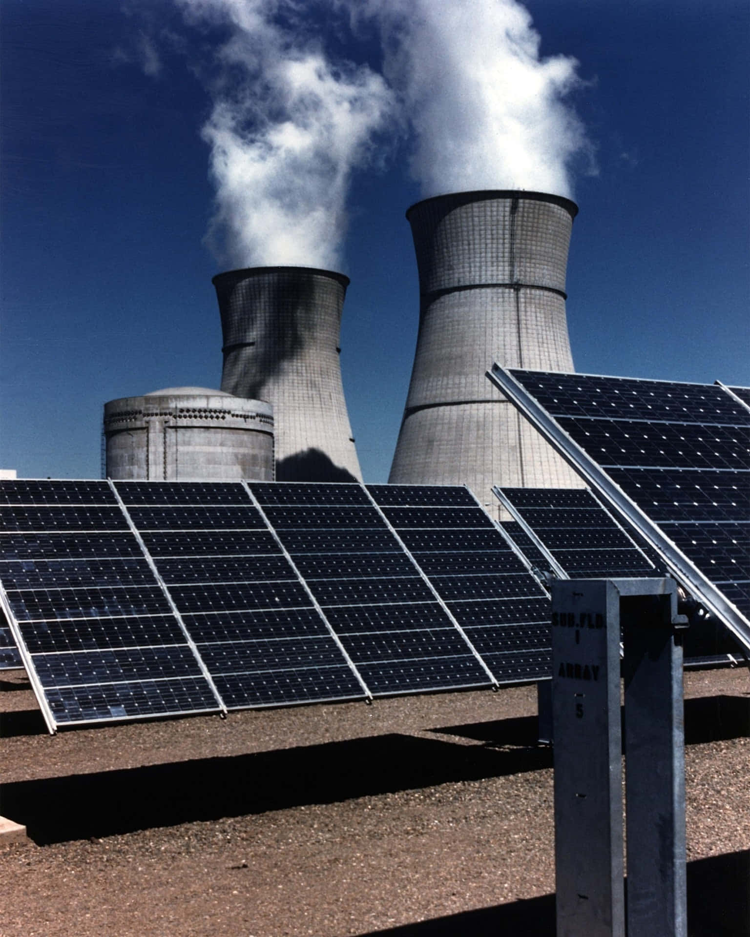 電力 桌布 壁紙 — 前景的太陽能板與高聳的核電廠冷卻塔形成對比，冷卻塔在晴朗的藍天下釋放出蒸汽。