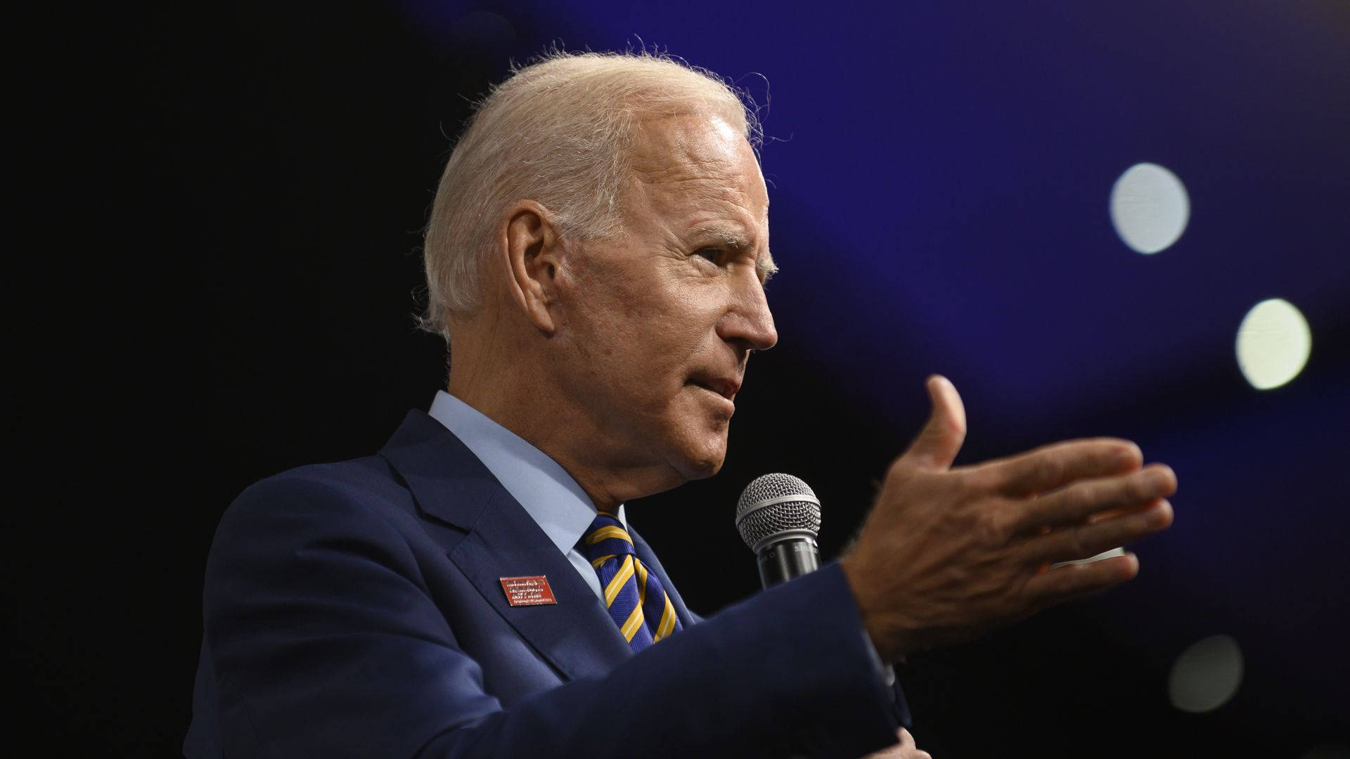 Biden 桌布 壁紙 — 前總統喬·拜登對著麥克風說話，表情嚴肅，背景是帶有散景燈光的深藍色模糊背景。