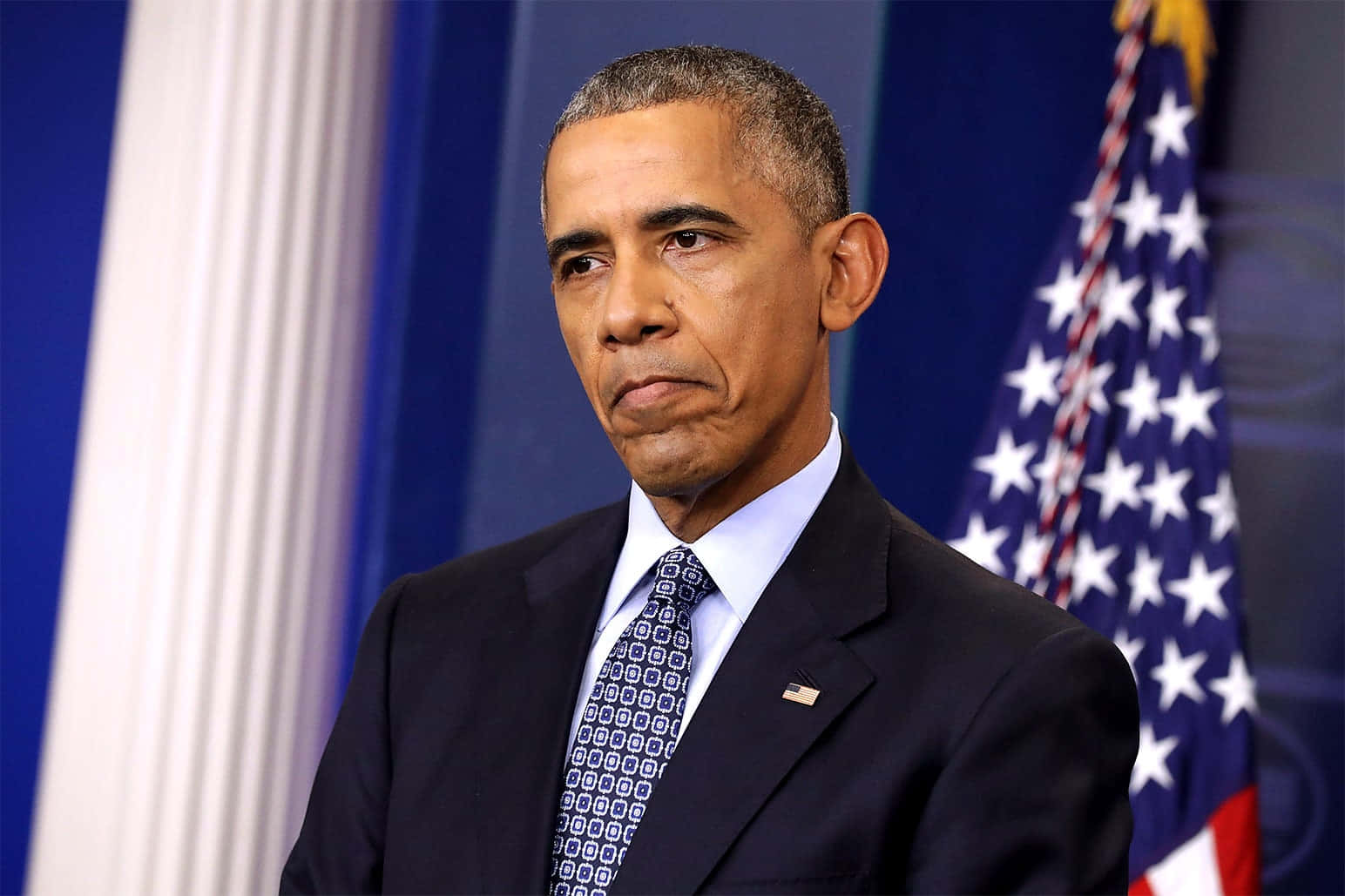 Barack Obama 桌布 壁紙 — 前總統巴拉克·歐巴馬身穿深色西裝和圖案領帶，莊嚴地站在美國國旗和藍色窗簾前。
