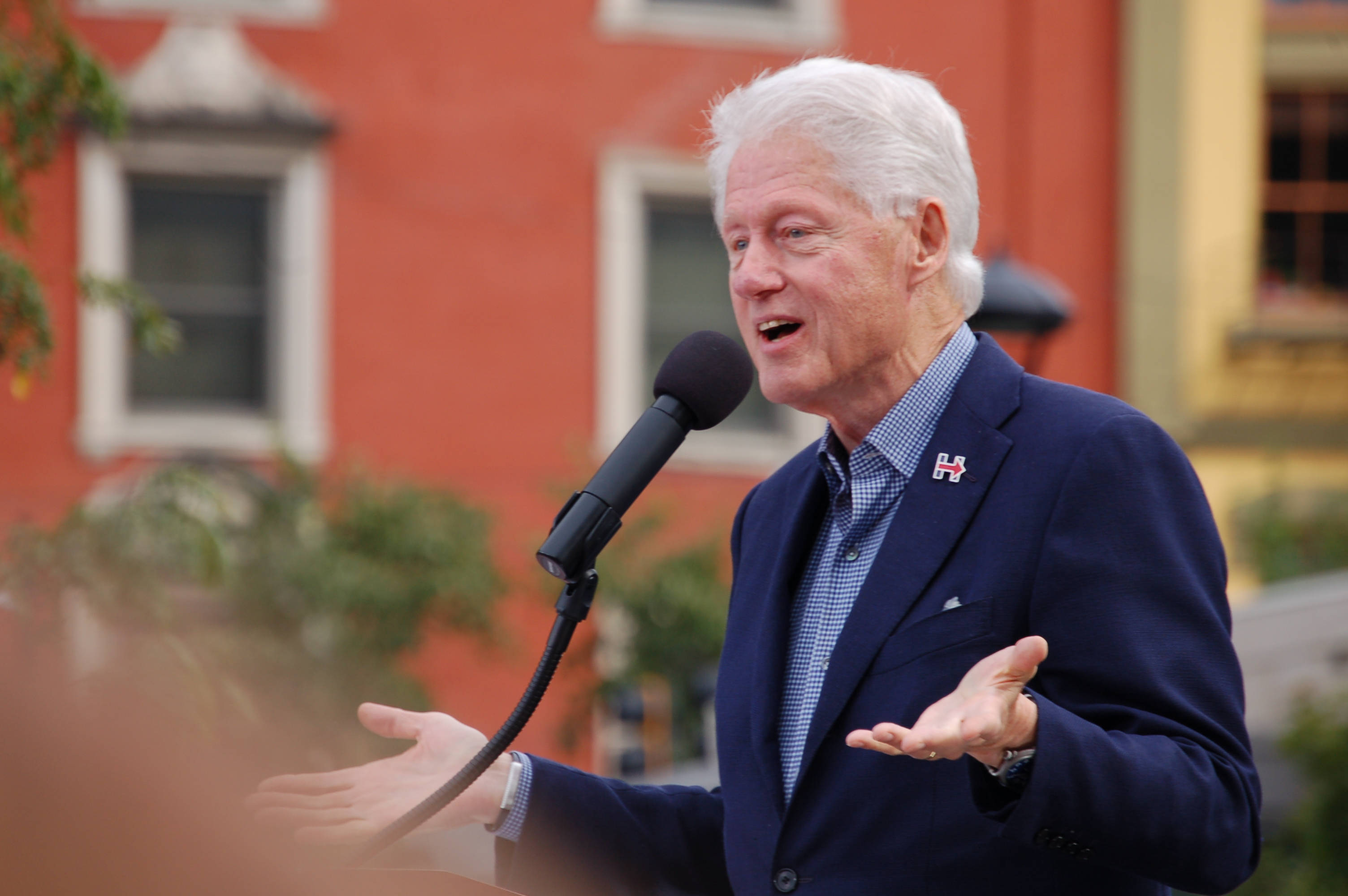 bill clinton 桌布 壁紙 — 前總統比爾·克林頓在戶外對著麥克風熱情演講，身後是一棟紅磚建築。