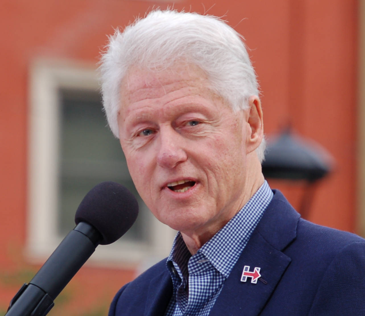bill clinton 桌布 壁紙 — 前總統比爾·克林頓在戶外面對麥克風發言，背景為紅磚建築。