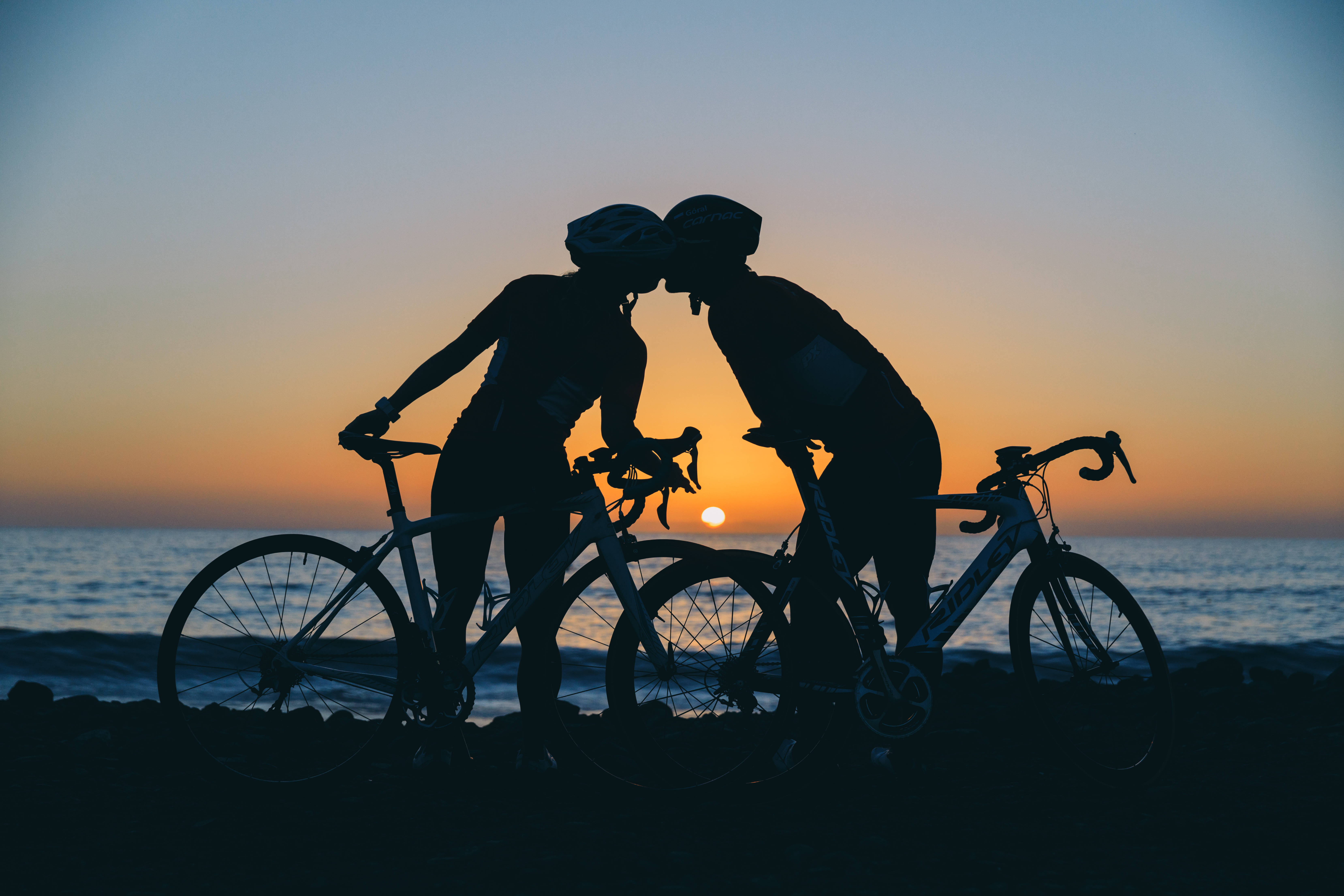 Cycling 桌布 壁紙 — 剪影：一對情侶在沙灘上日落時分接吻，旁邊有腳踏車，背景是橙色天空。