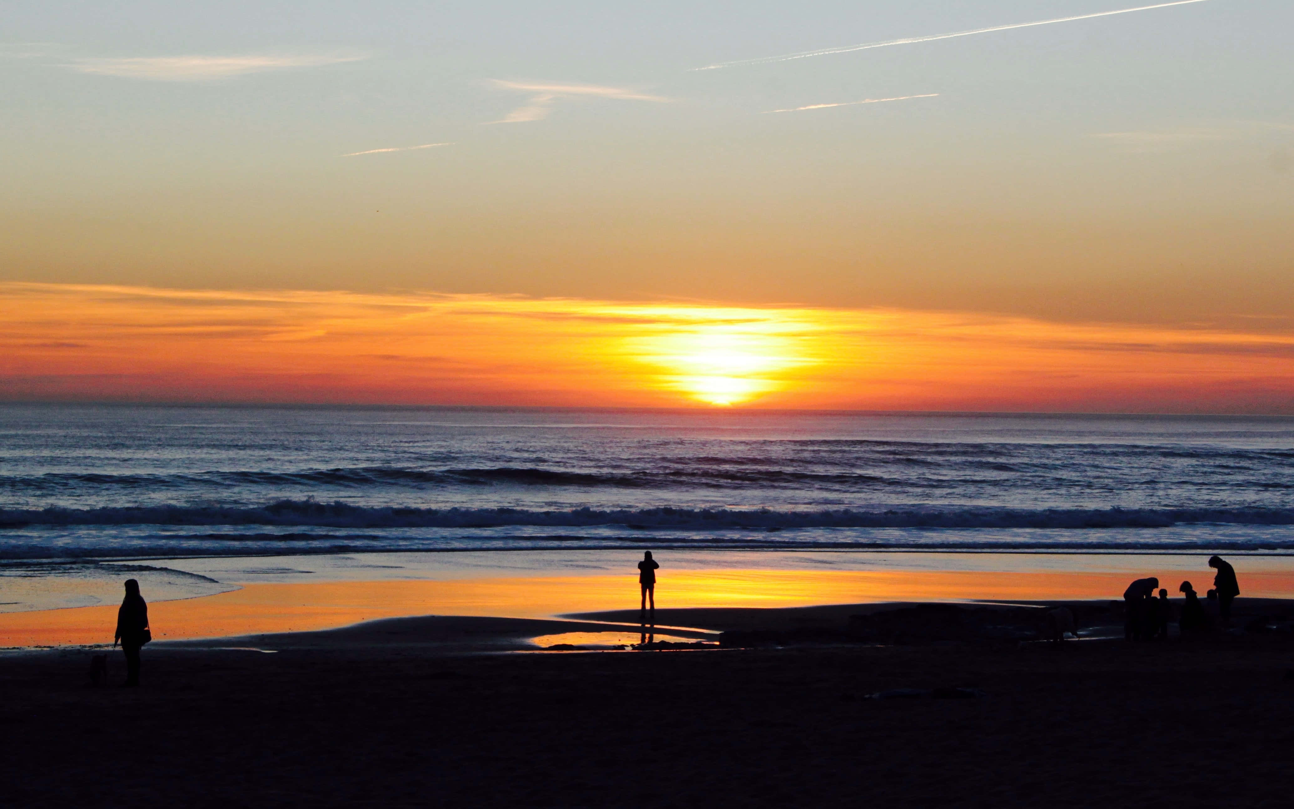 Sunset Beach 桌布 壁紙 — 剪影人物在海灘上漫步和聚集，夕陽以火熱的橙色和金色沉入海洋。