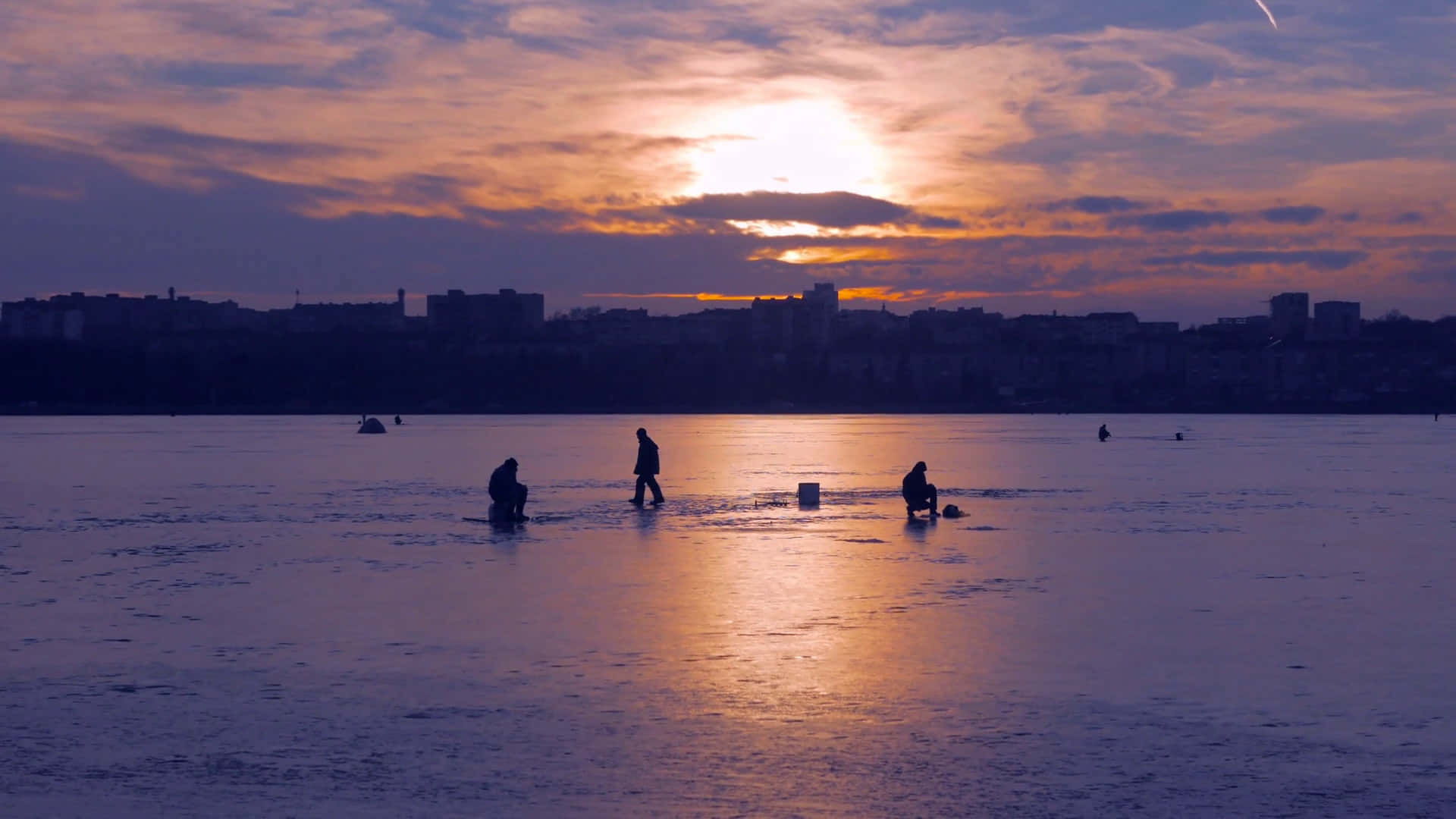 Ice Fishing 桌布 壁紙 — 剪影人物在結冰的湖面上釣魚，夕陽以橙色和紫色渲染天空。