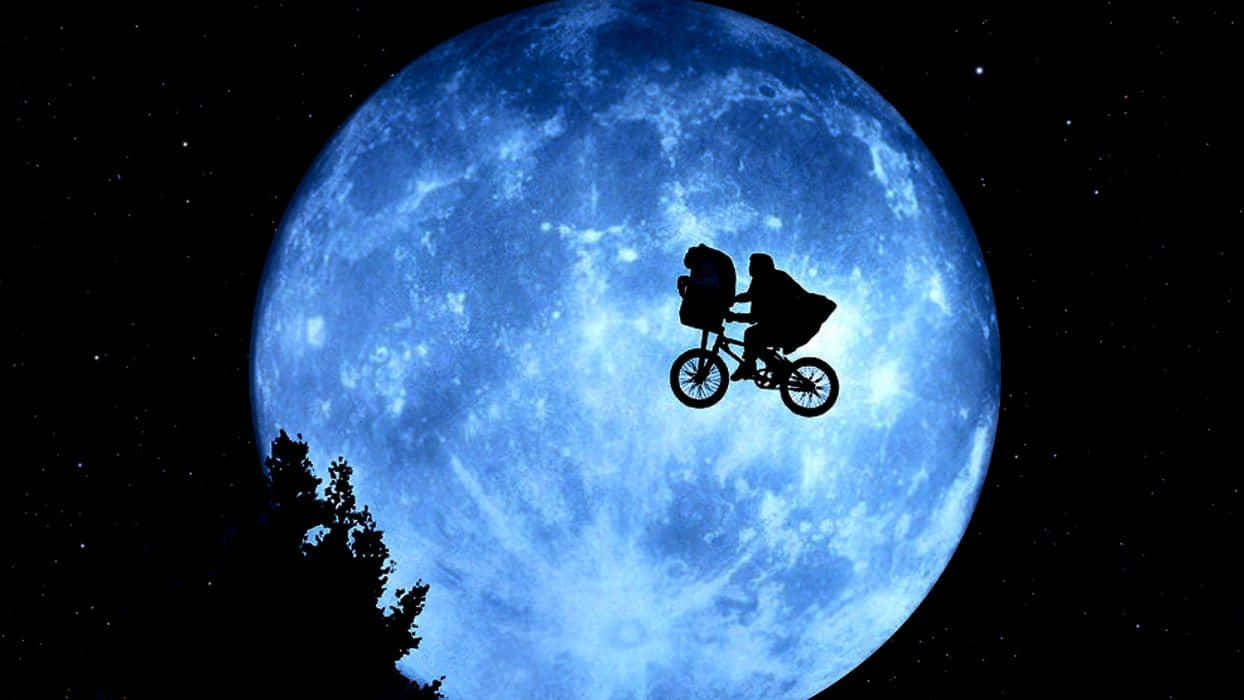 Et 桌布 壁紙 — 剪影人物在繁星點點的夜空下，騎著腳踏車，背景是一輪巨大的藍色月亮，樹梢清晰可見。