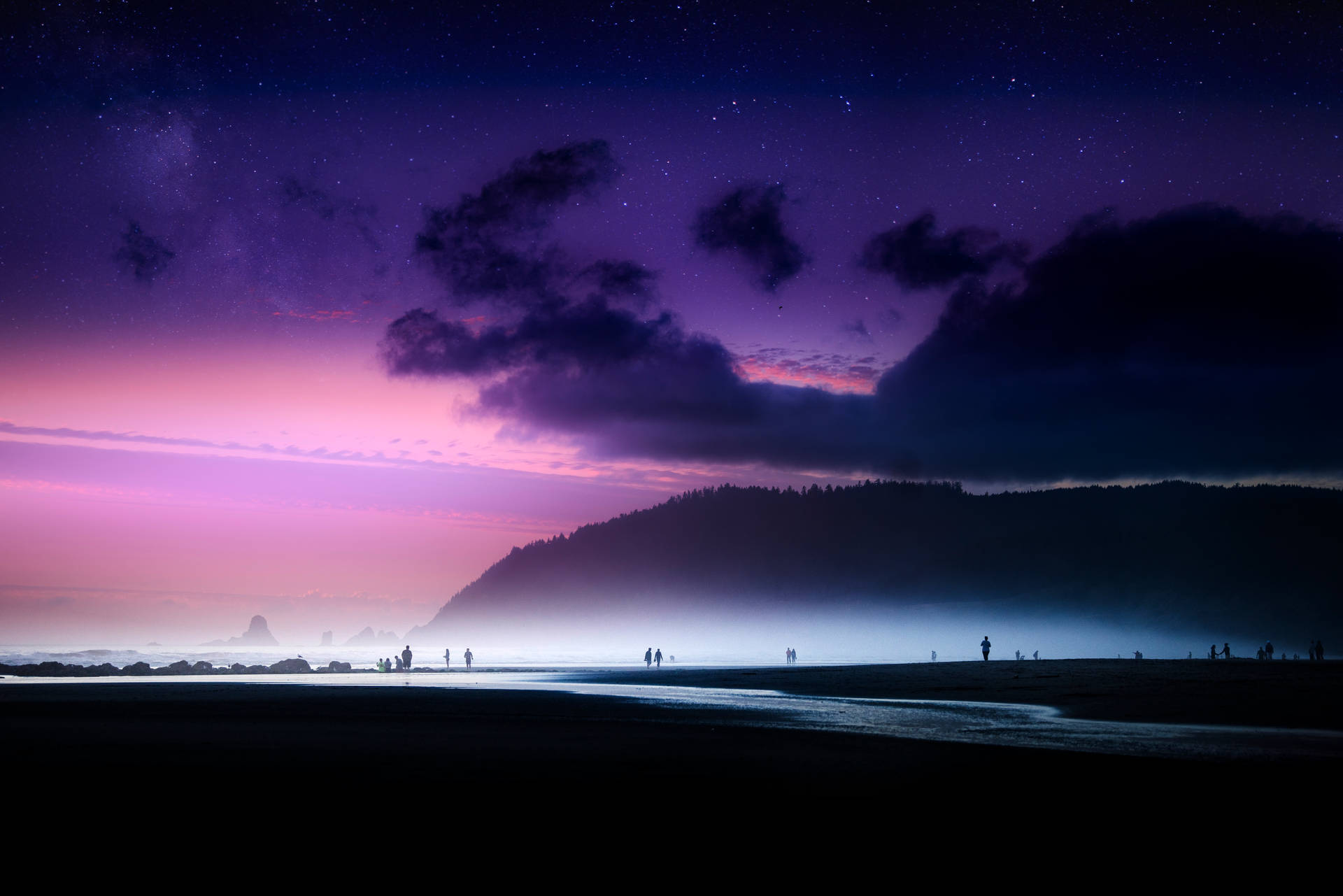 人們在暮色中於紫色星空下漫步在霧氣瀰漫的海灘上。
