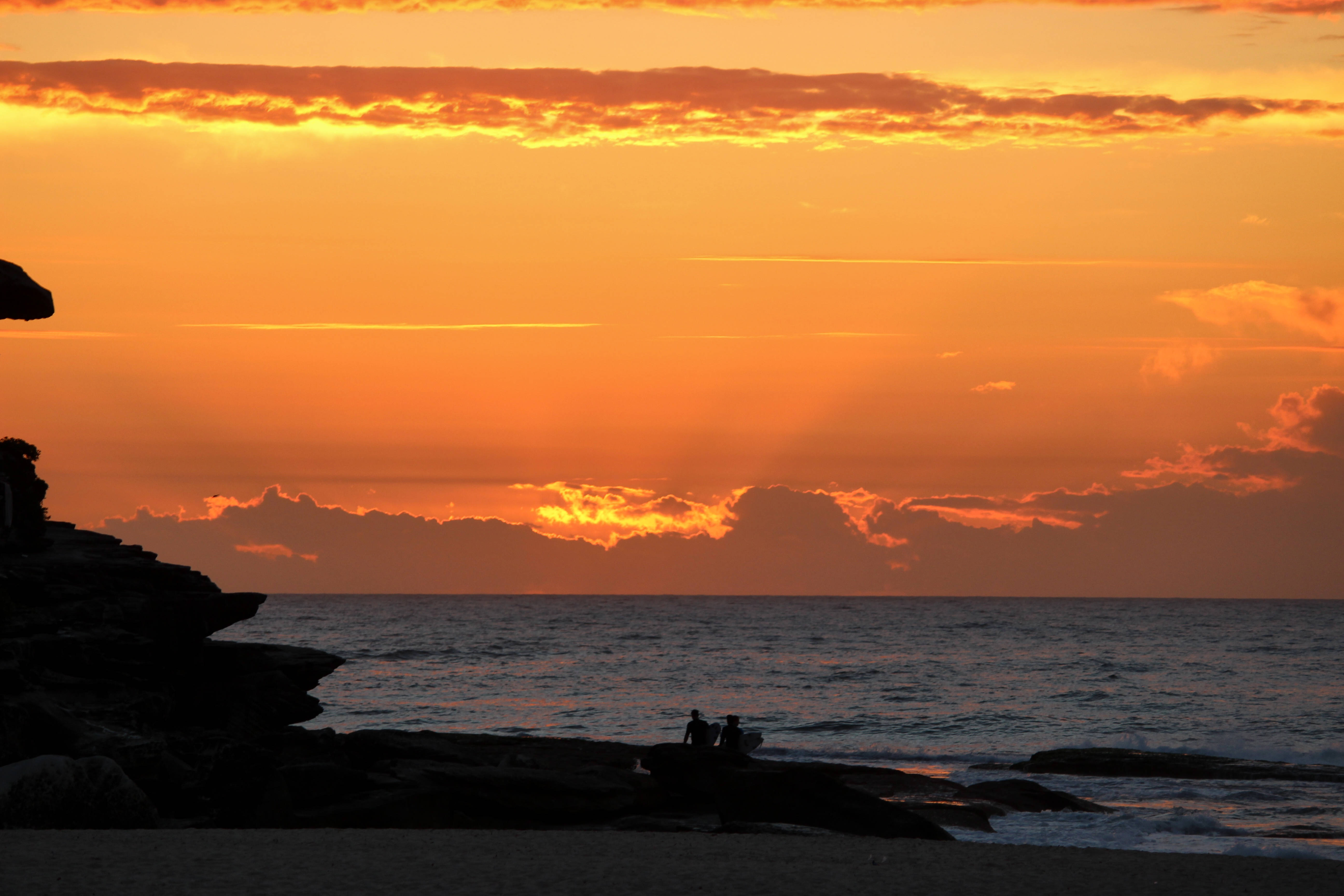Friendship 桌布 壁紙 — 剪影人物坐在岩石海岸上，火橙色的夕陽將雲彩和海洋染成溫暖的光芒。