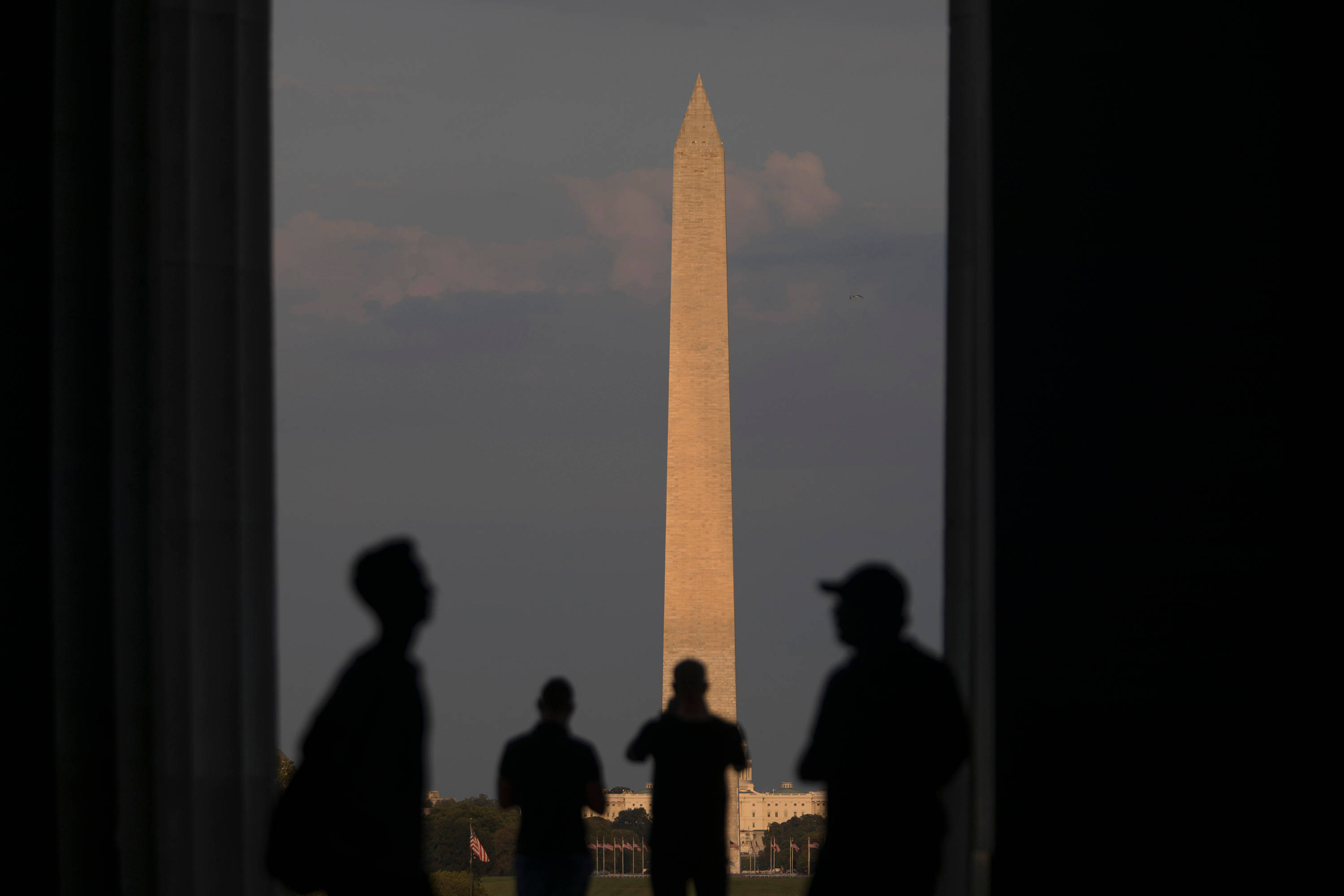 Washington 桌布 壁紙 — 剪影人物走過一個宏偉的門口，框住沐浴在金色日落光芒中的華盛頓紀念碑。