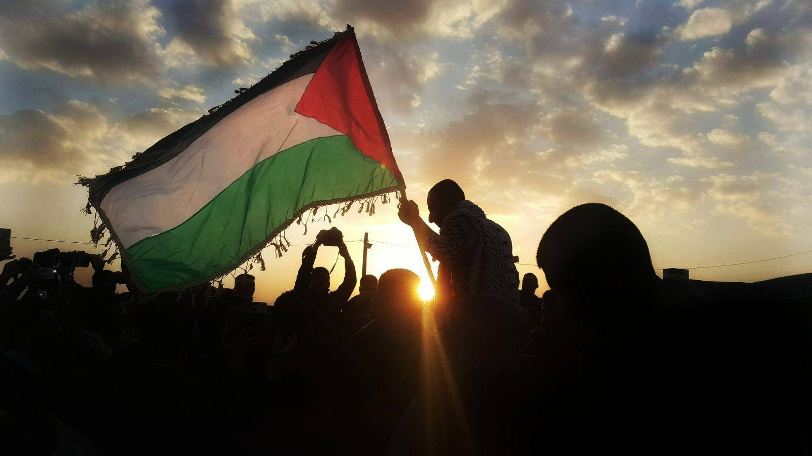 Palestine 桌布 壁紙 — 剪影人群高舉巴勒斯坦旗幟，背景是金色夕陽天空和戲劇性的雲彩。