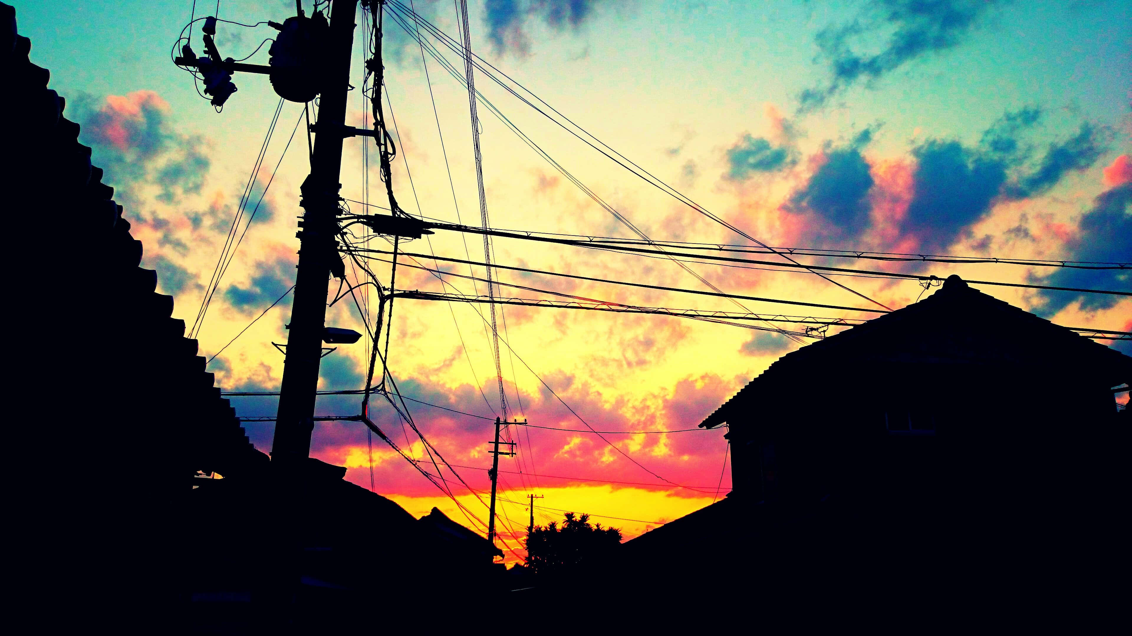 Anime Sky 桌布 壁紙 — 剪影屋頂和電線框住了充滿活力的日落天空，帶有火熱的橙色、粉紅色和藍綠色雲彩。