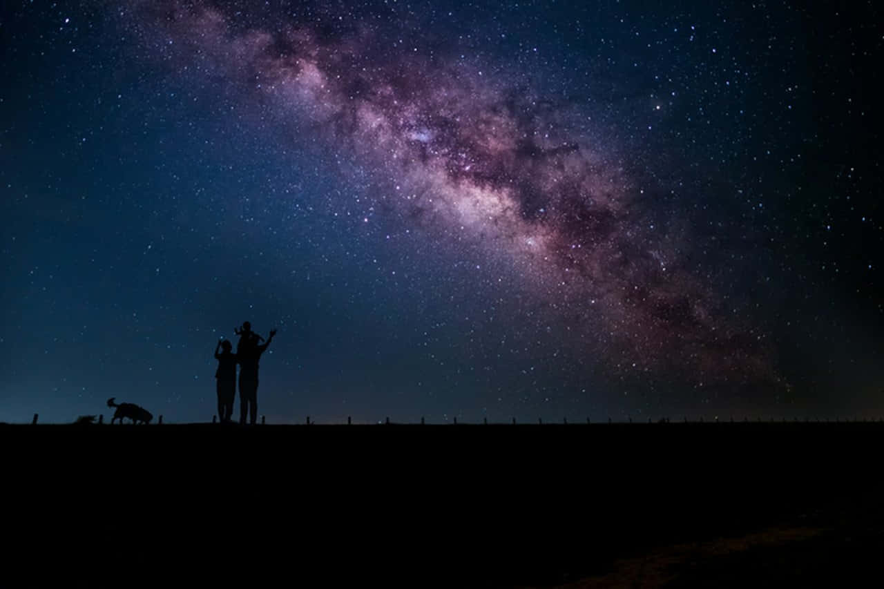 Beautiful Galaxy 桌布 壁紙 — 剪影情侶和狗仰望橫跨繁星夜空、色彩鮮豔的紫藍色銀河。