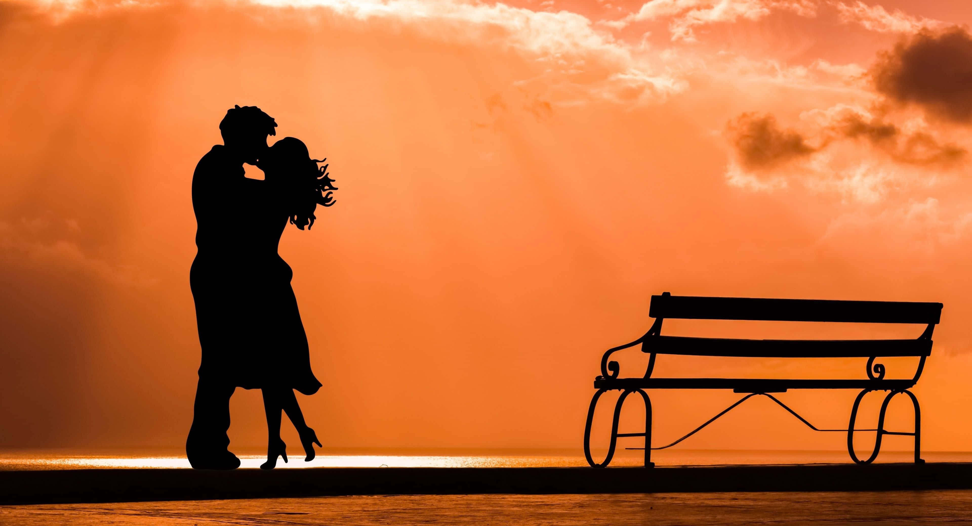 couple love 桌布 壁紙 — 剪影情侶在日落時分於空長椅旁熱情擁抱，背景是火紅的橙色天空。
