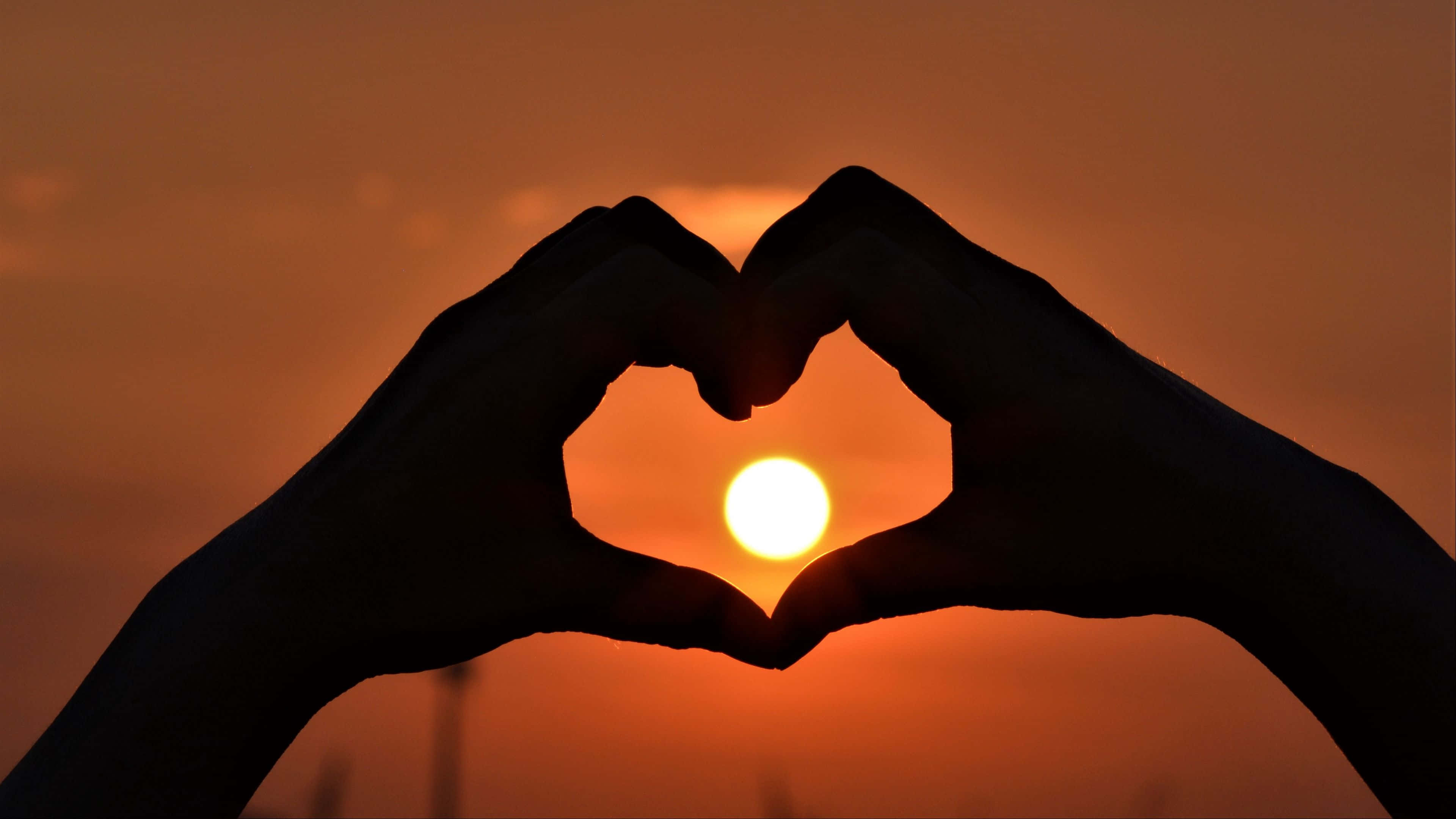 4K Love 桌布 壁紙 — 剪影手形成心形，框住溫暖橙色天空中的發光夕陽。