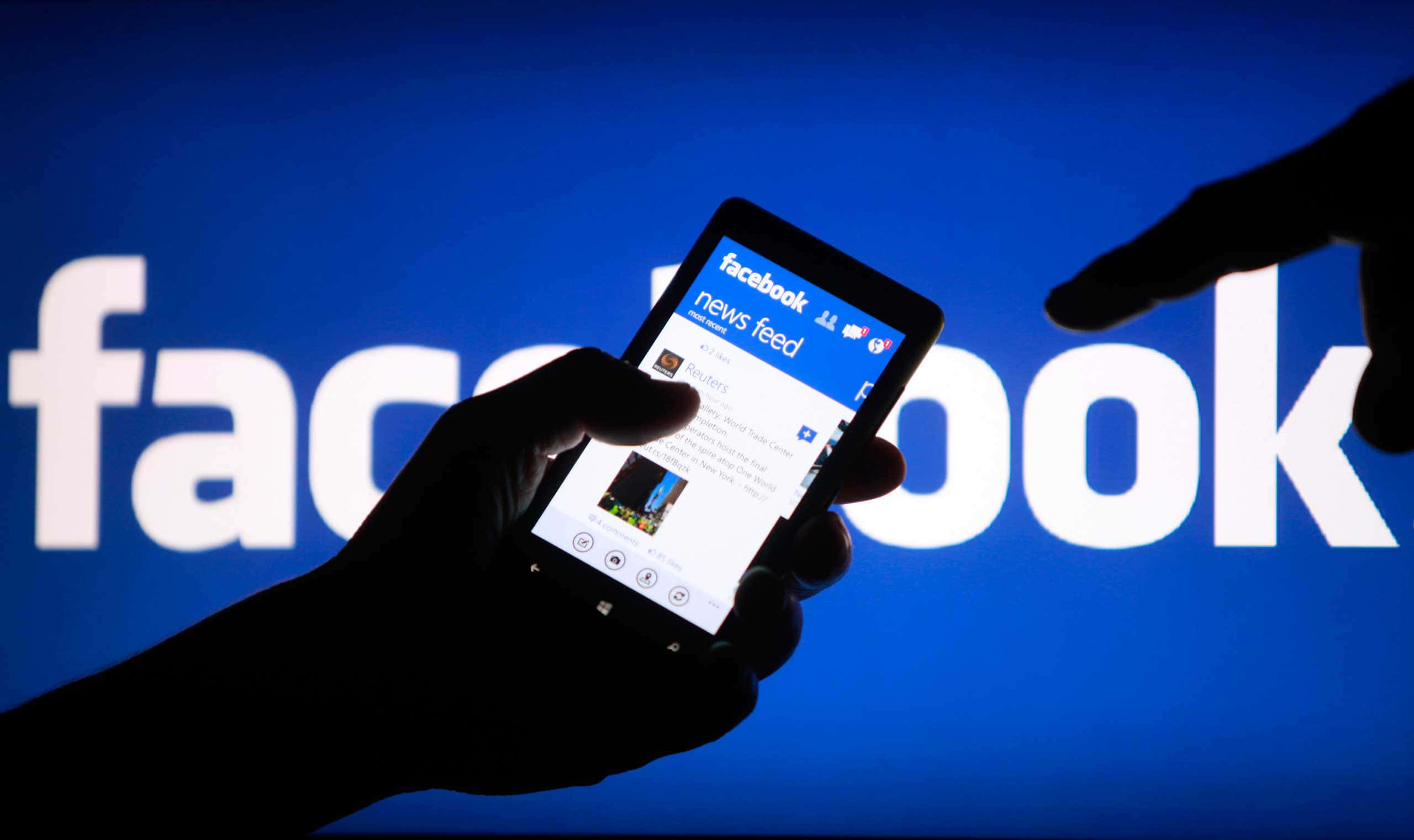 Facebook 桌布 壁紙 — 剪影手部拿著智慧型手機，顯示Facebook新聞動態，背景是發光的藍色Facebook標誌。