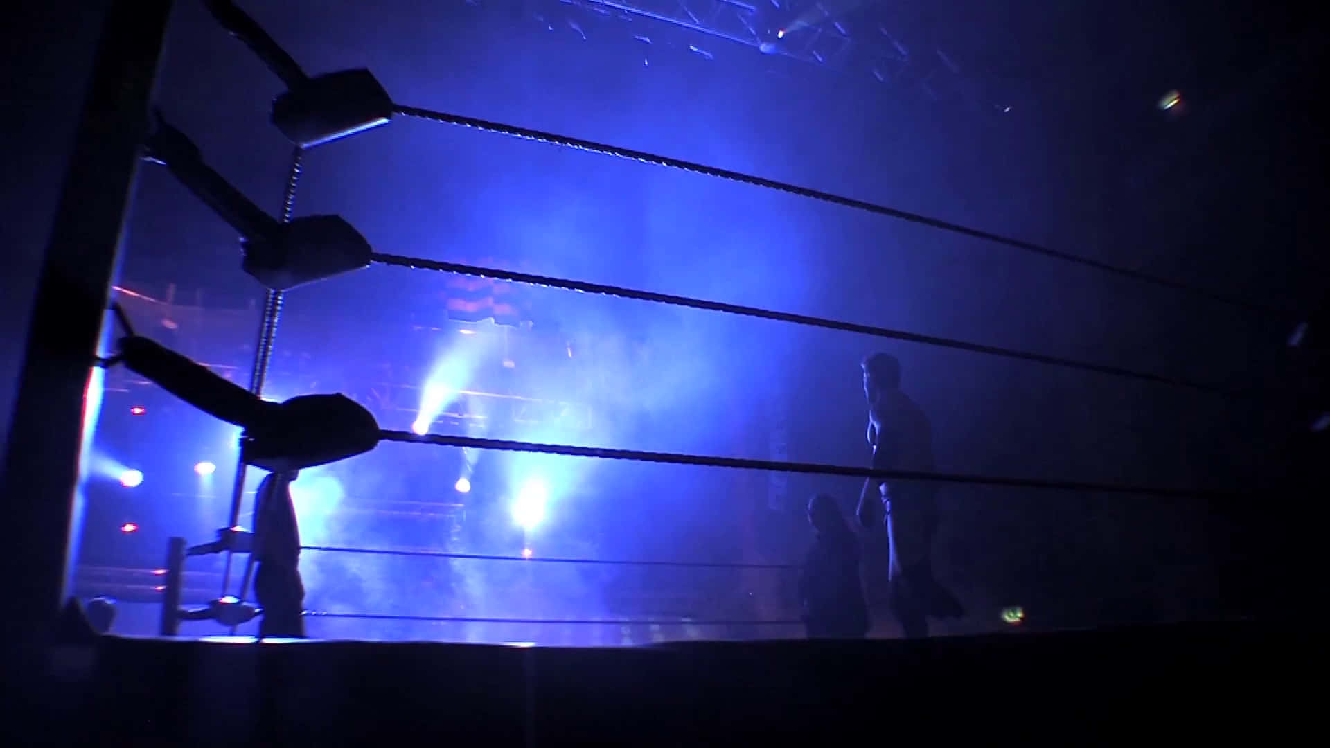 Wrestling Ring 桌布 壁紙 — 剪影摔角手站在昏暗的擂台上，沐浴在電藍色舞台燈光和煙霧中。