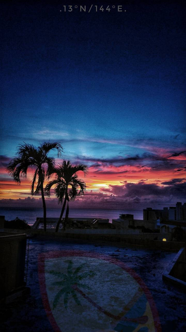 Guam 桌布 壁紙 — 剪影棕櫚樹框出從屋頂（帶有彩繪盾牌）看到的海洋和城市天際線上 vibrant 的日落。