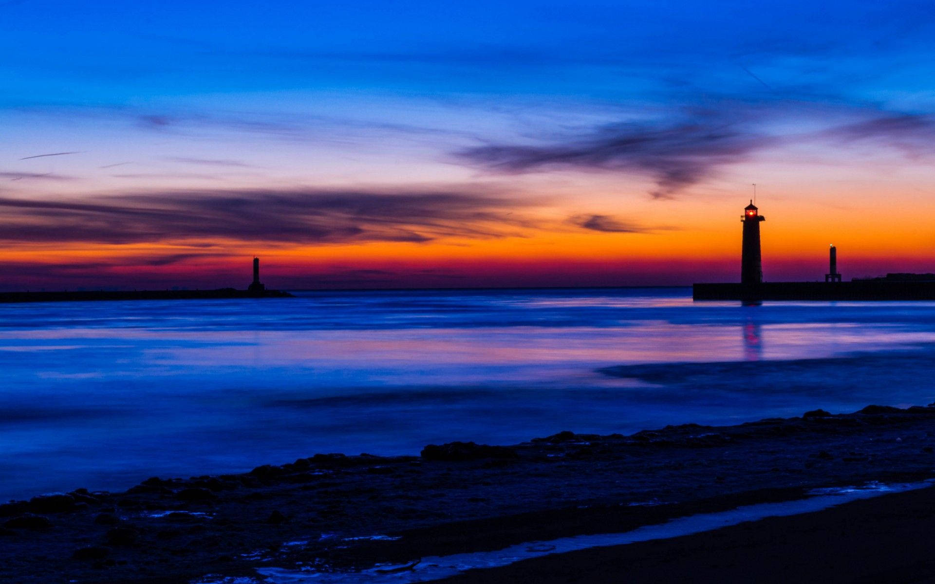 Lighthouse 桌布 壁紙 — 剪影燈塔矗立在火紅的日落天空下，倒映在平靜的藍色海水中，前景是岩石海岸。