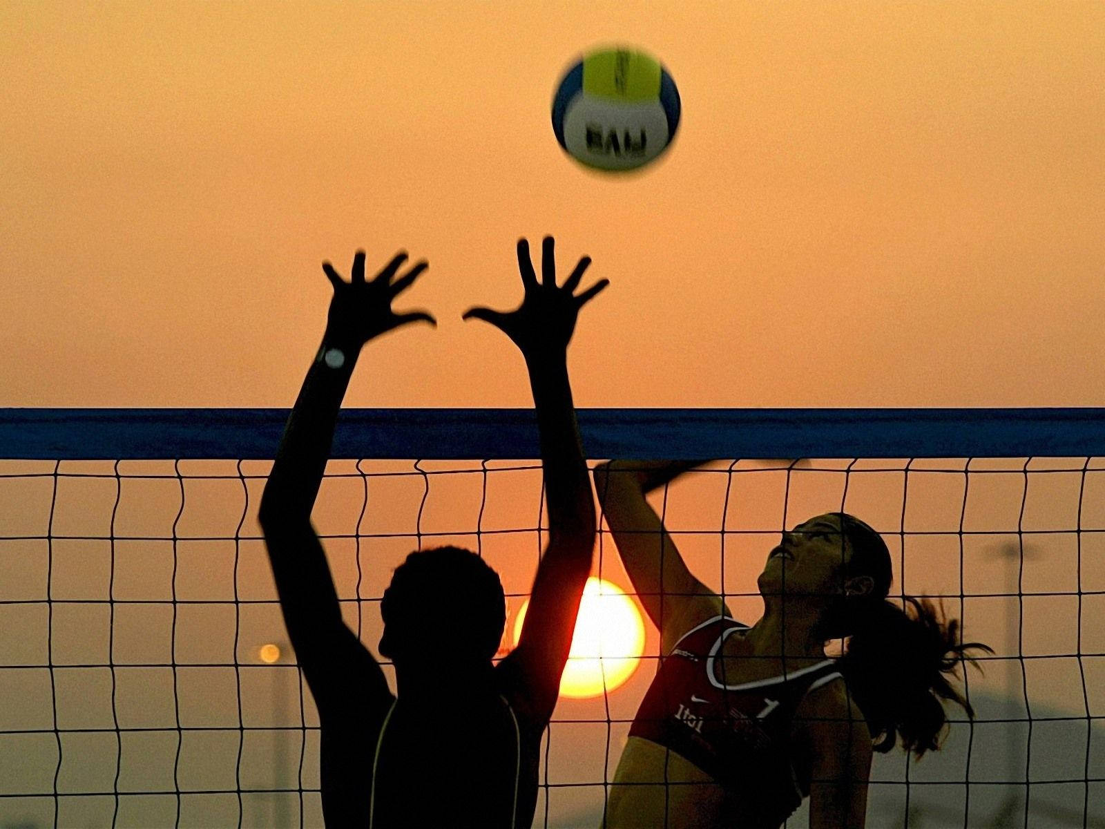 Volleyball 桌布 壁紙 — 剪影的排球運動員在日落時跳躍，球越過球網，映襯著發光的橙色天空。