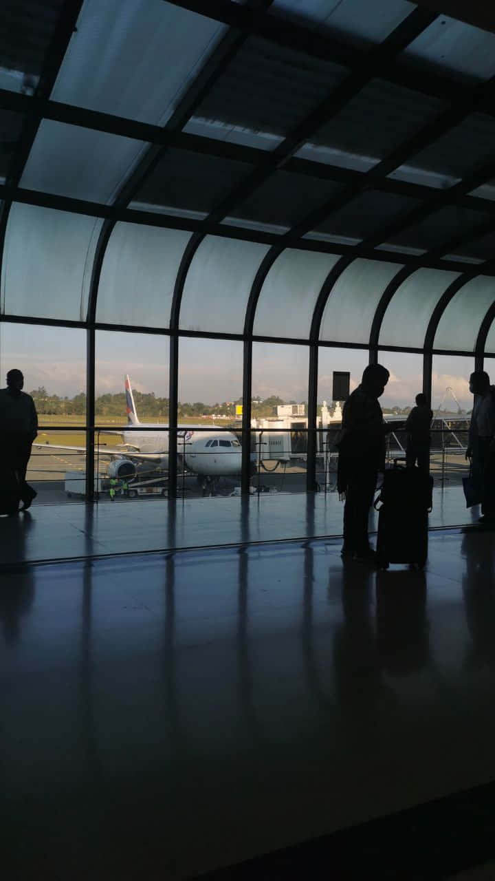 Airport 桌布 壁紙 — 剪影的旅客帶著行李站在現代機場航站樓內，透過彎曲的玻璃窗凝視著停放的飛機。