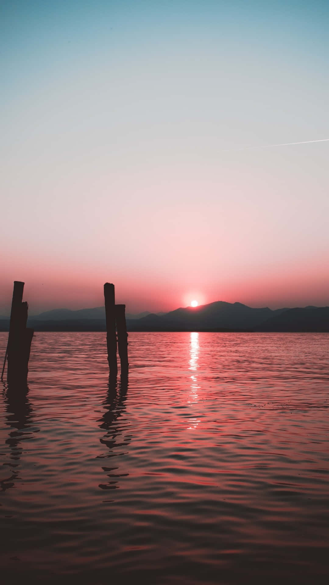 Calming iPhone 桌布 壁紙 — 剪影的柱子矗立在平靜的水中，鮮豔的粉紅色夕陽在遠山之上發光。