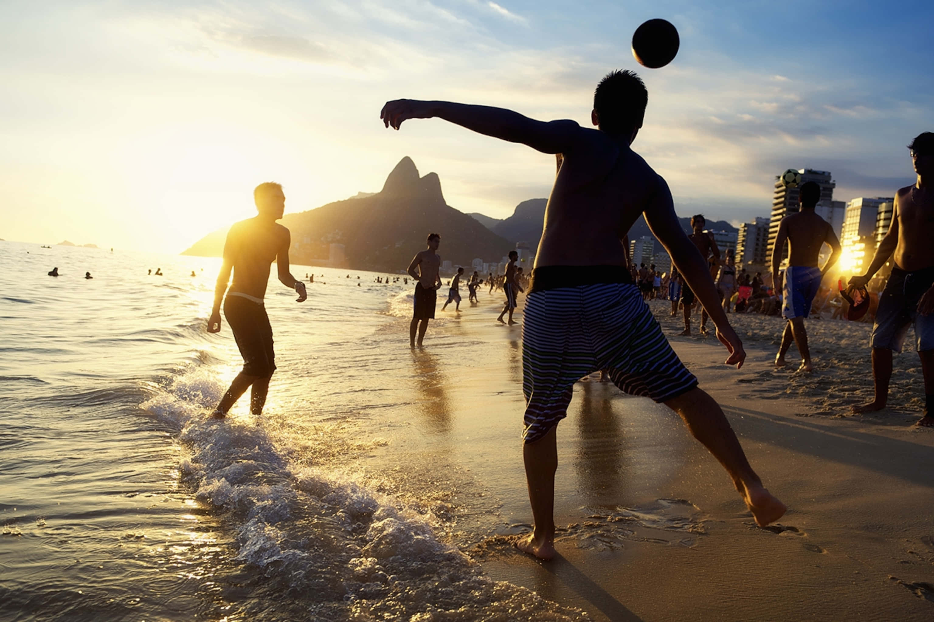 Brazilian Beach 桌布 壁紙 — 剪影的海灘遊客在日落時踢足球，海浪拍打，背景是標誌性的山峰。