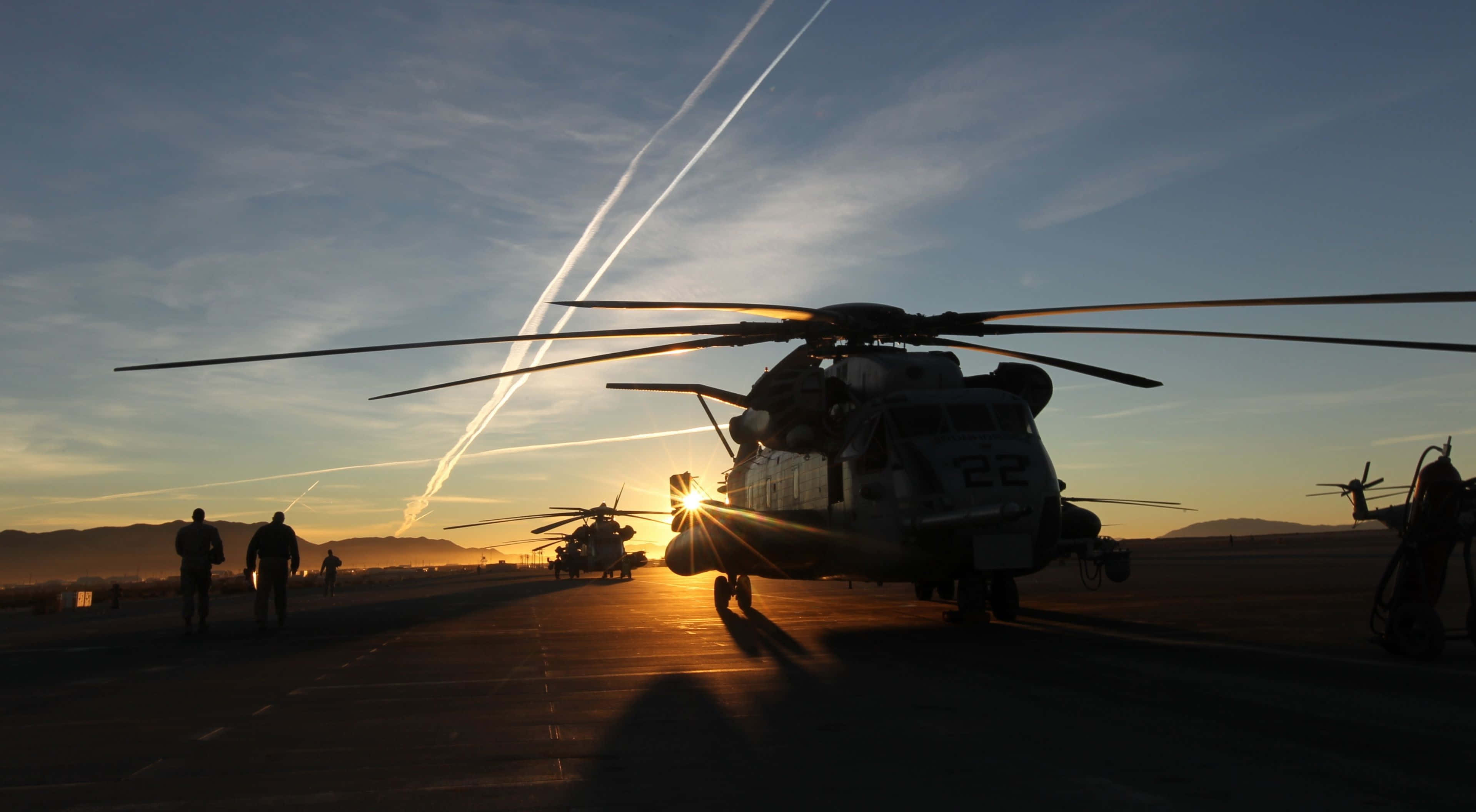 4K Helicopters 桌布 壁紙 — 剪影的軍用直升機在日落時分停在停機坪上，機組人員在附近走動，凝結尾跡劃過天空。