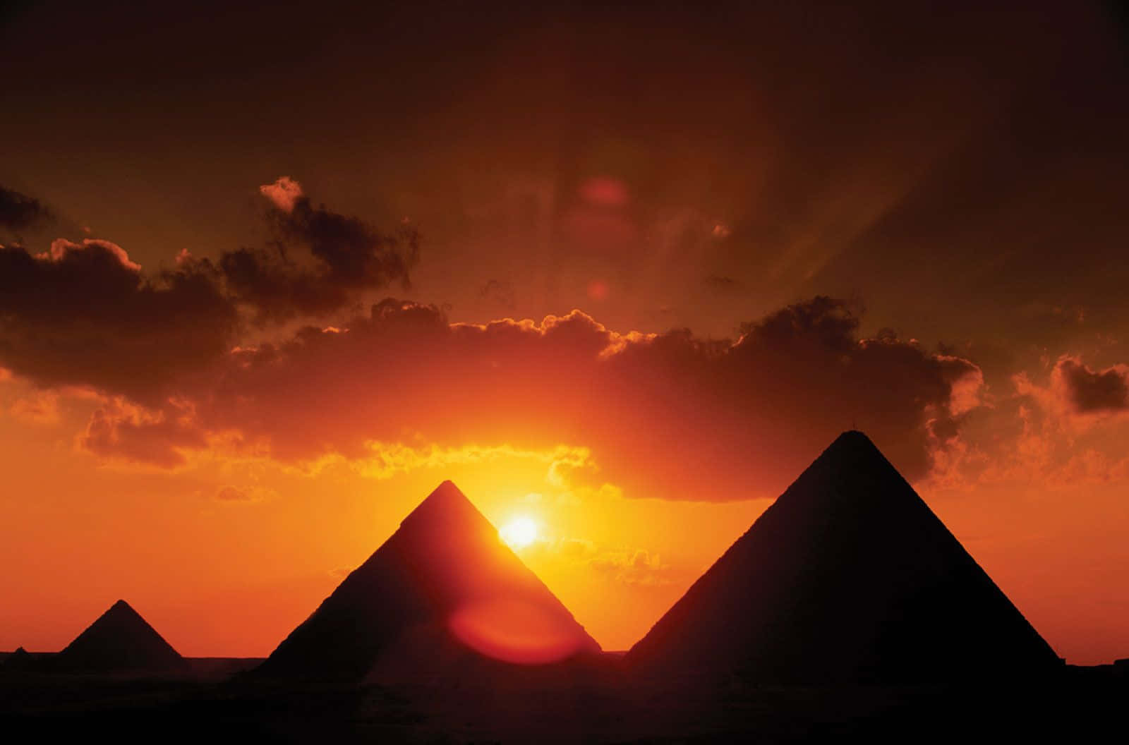 沙漠 桌布 壁紙 — 剪影的金字塔在火紅的日落天空下聳立，伴隨著發光的雲彩和燦爛的陽光。