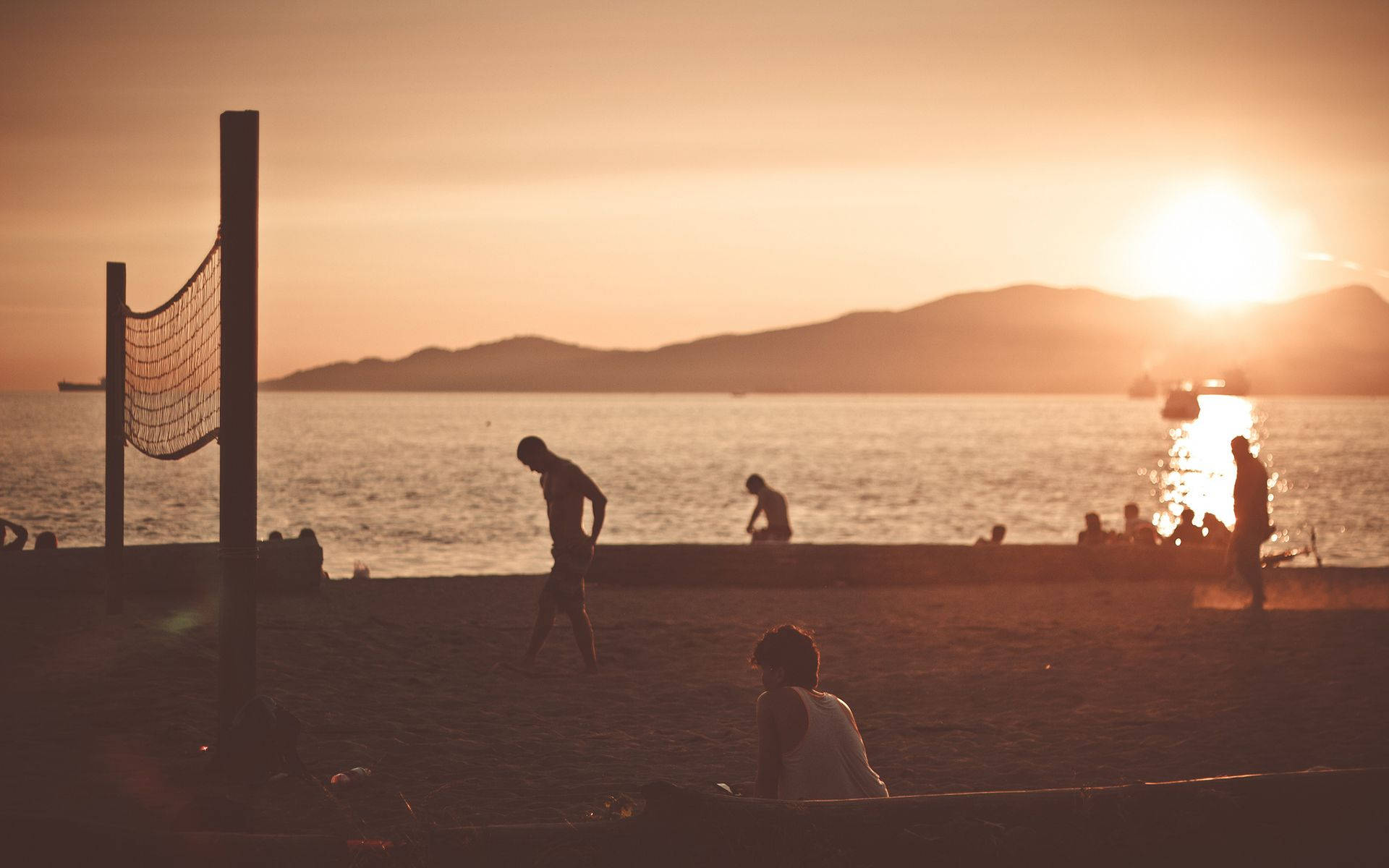 海灘 桌布 壁紙 — 剪影般的海灘遊客在平靜的水面和遠處山丘上夕陽西下時打排球。