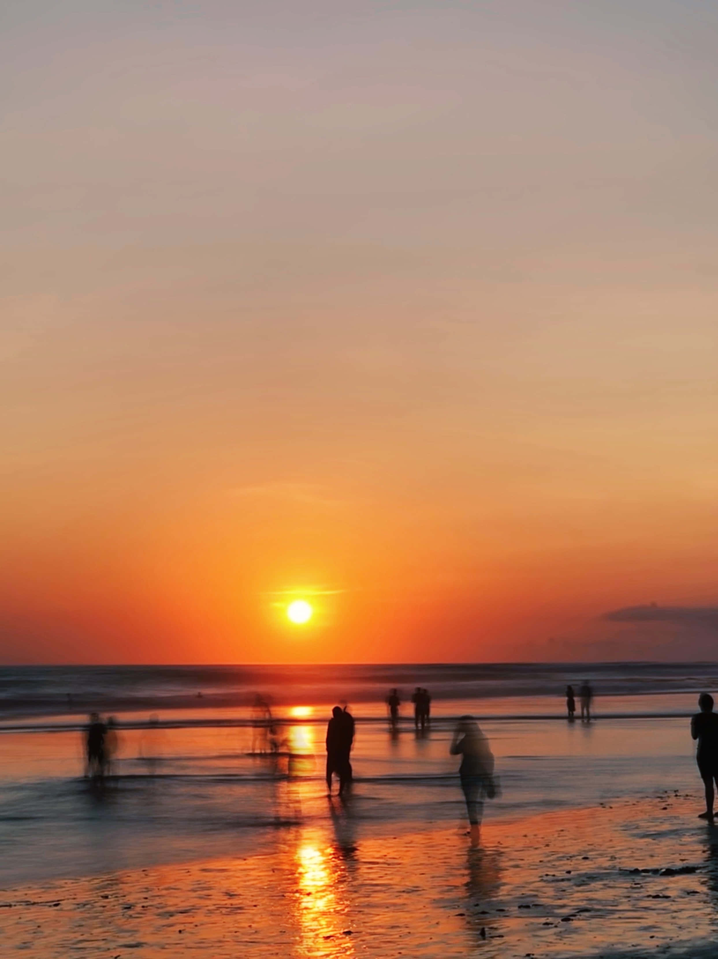 Sunset Beach 桌布 壁紙 — 剪影般的海灘遊客在濕沙上漫步，火紅的夕陽在平靜的海面上落下。