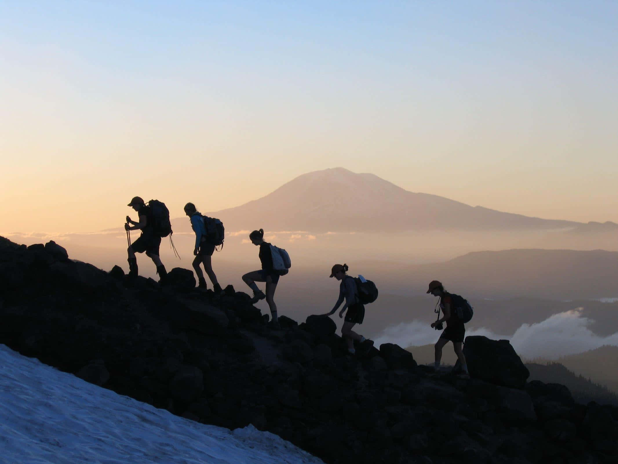 Hiking 桌布 壁紙 — 剪影般的登山者在黎明時分攀登積雪的山脊，身後升起霧氣繚繞的山峰。