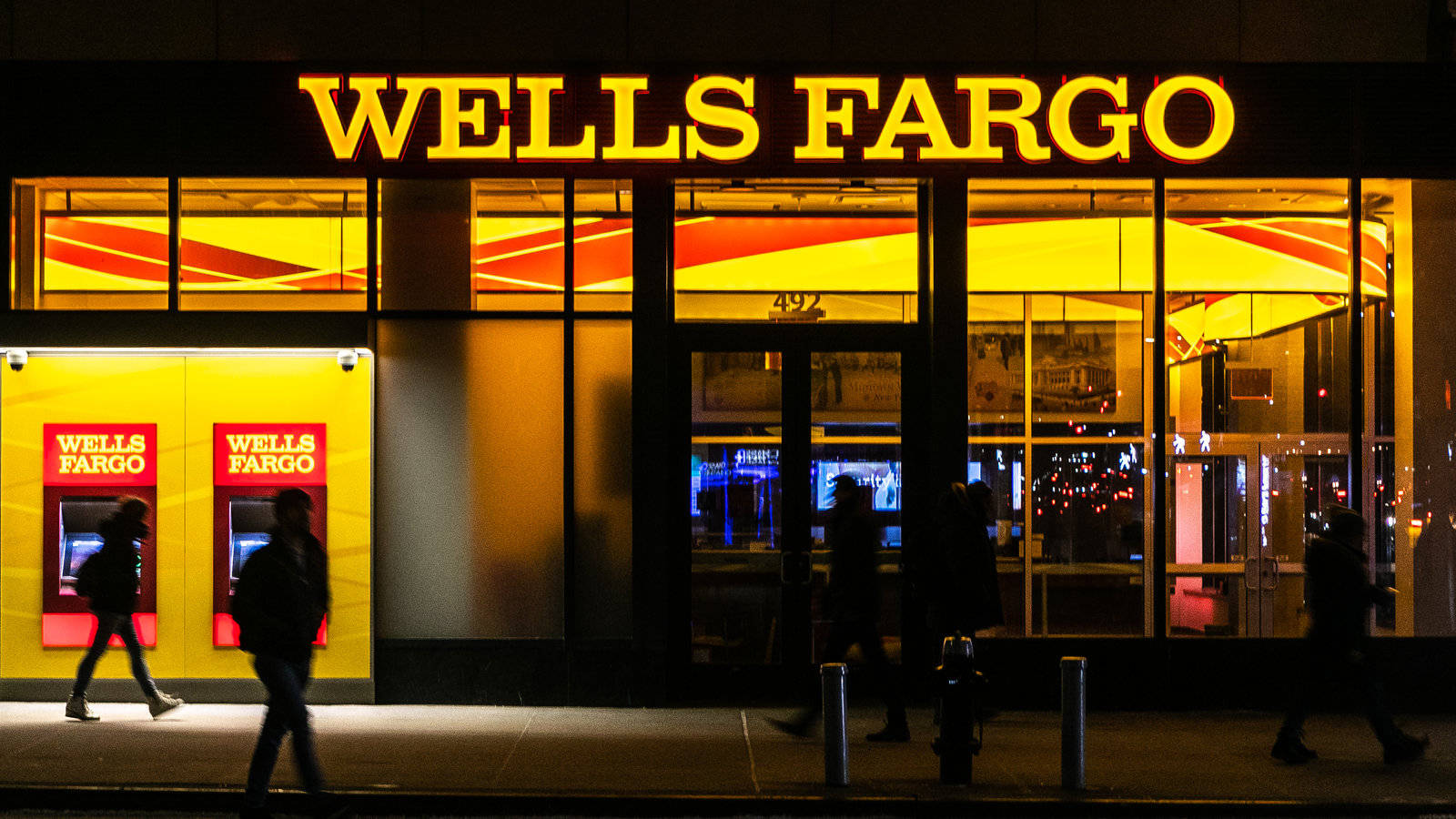 sign 桌布 壁紙 — 剪影般的行人夜晚經過燈火通明的 Wells Fargo 銀行，明亮的黃色標誌和玻璃窗反射城市燈光。