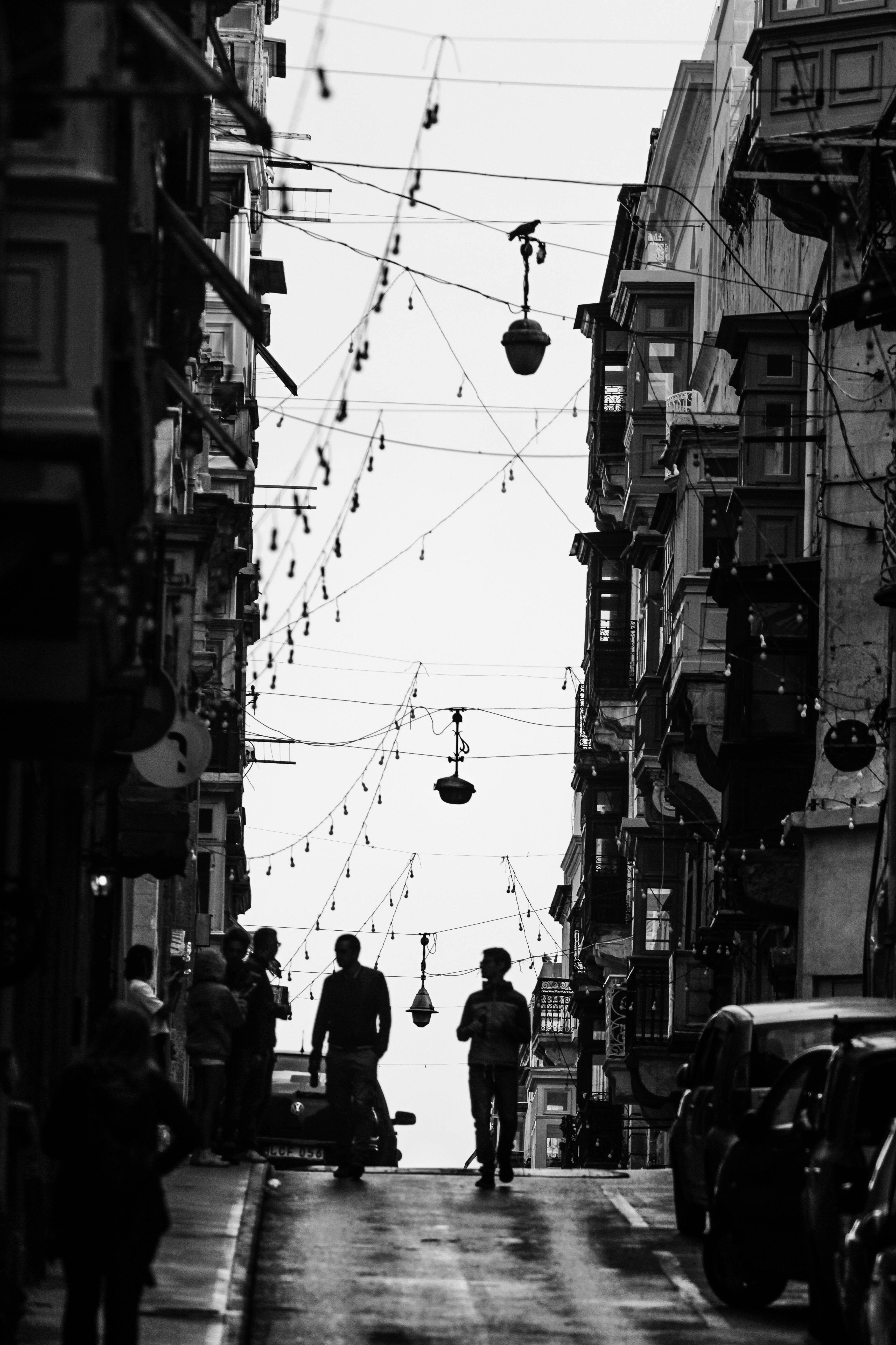 Malta 桌布 壁紙 — 剪影般的行人走在一條狹窄潮濕的街道上，兩旁是高樓大廈，天空被懸掛的燈籠和電線穿透。