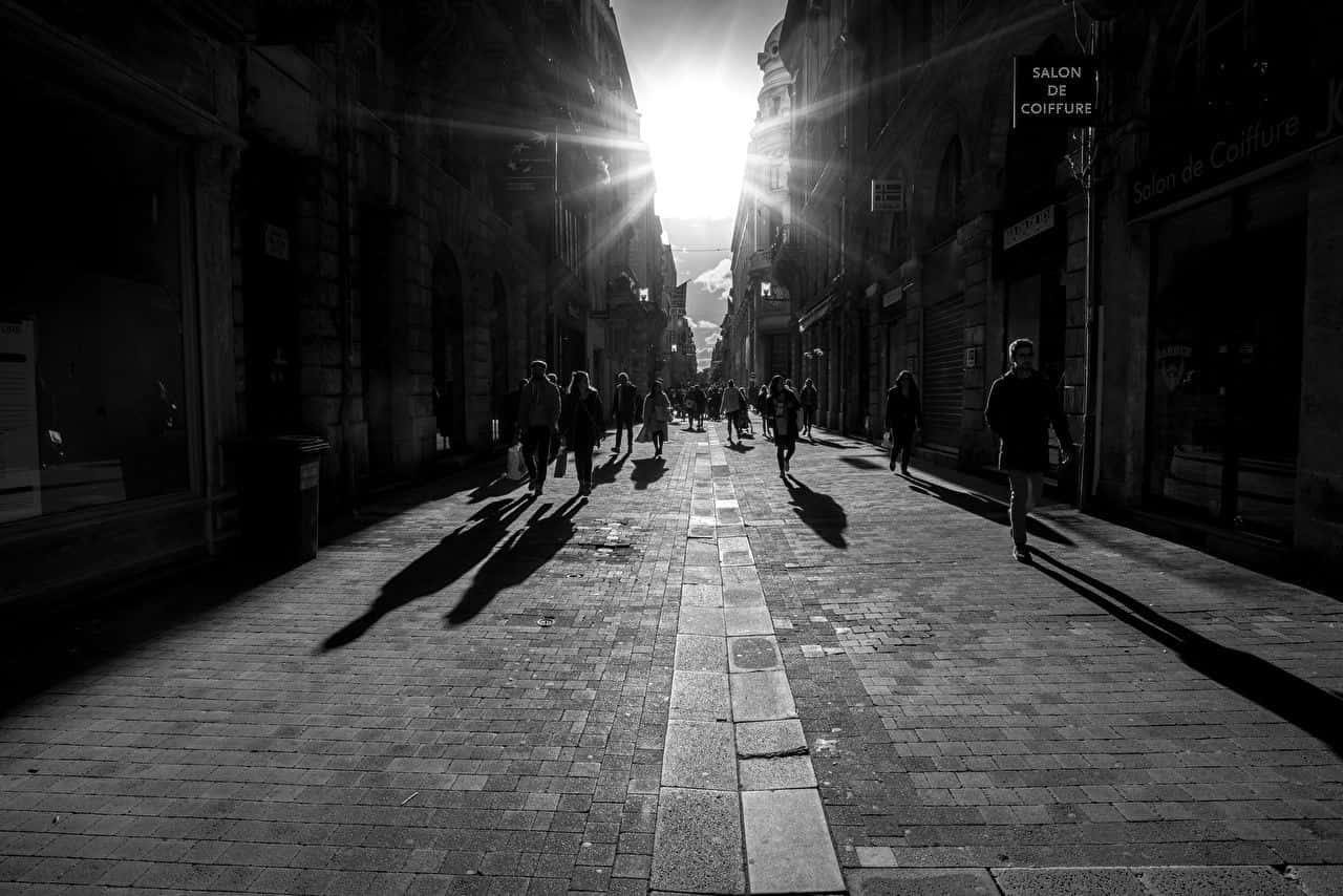 Black And White Street 桌布 壁紙 — 剪影般的行人走在一條狹窄的鵝卵石街道上，刺眼的陽光從頭頂的建築物之間照射進來。