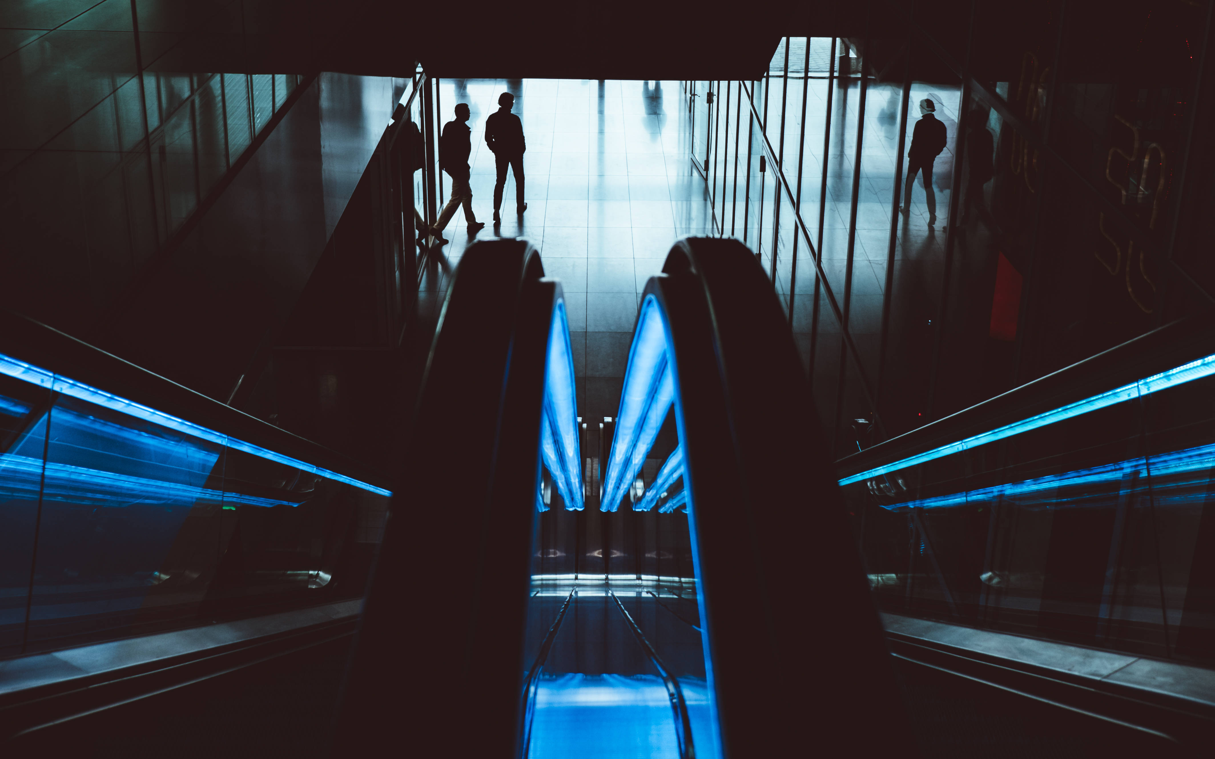 dark neon iPhone 桌布 壁紙 — 剪影般的通勤者在現代交通樞紐中，在冰冷的藍色電梯上上下下，沐浴在冷色調的人造光中。