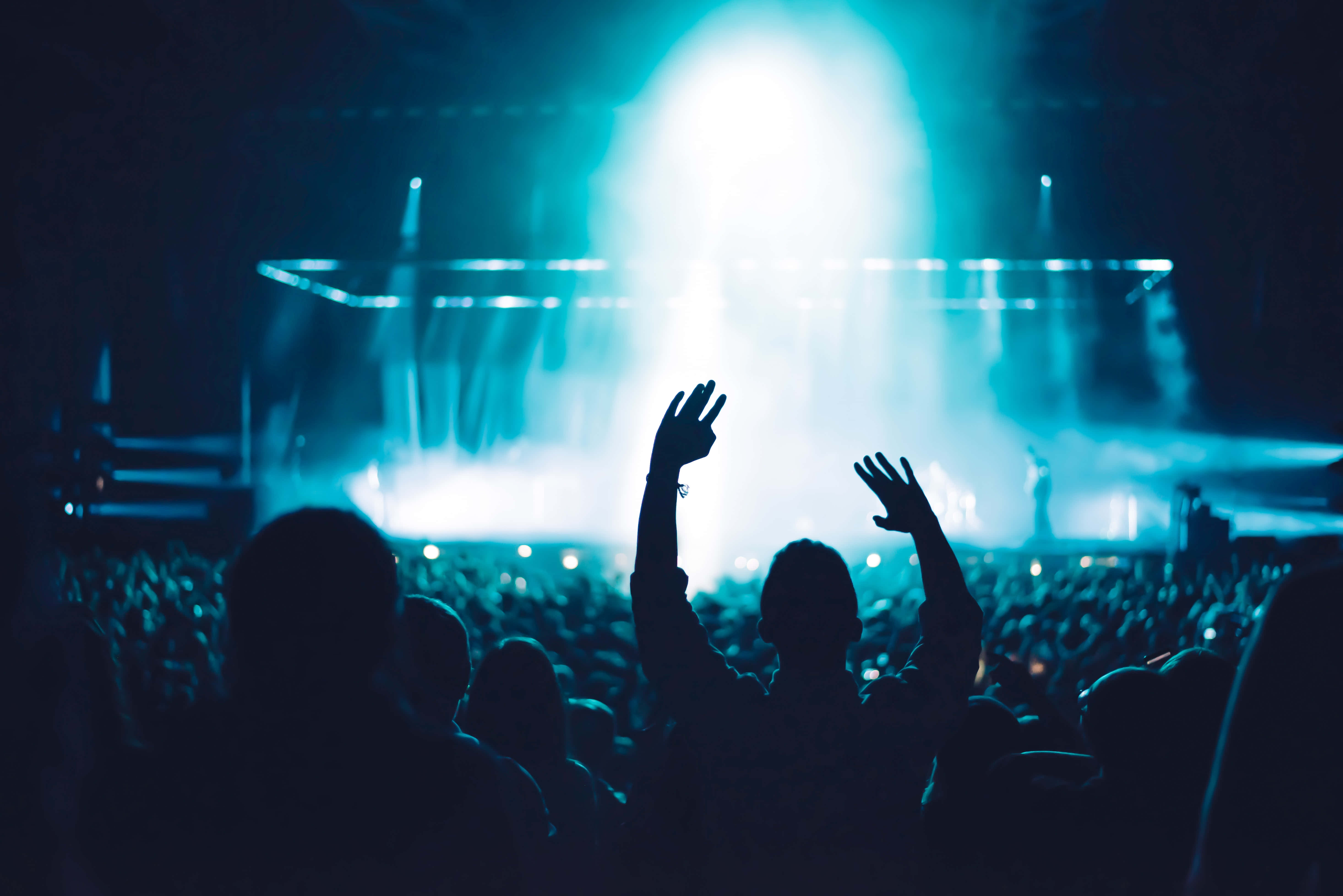music festival 桌布 壁紙 — 剪影般的音樂會觀眾向著沐浴在戲劇性聚光燈和煙霧中的發光藍色舞台舉起雙手。