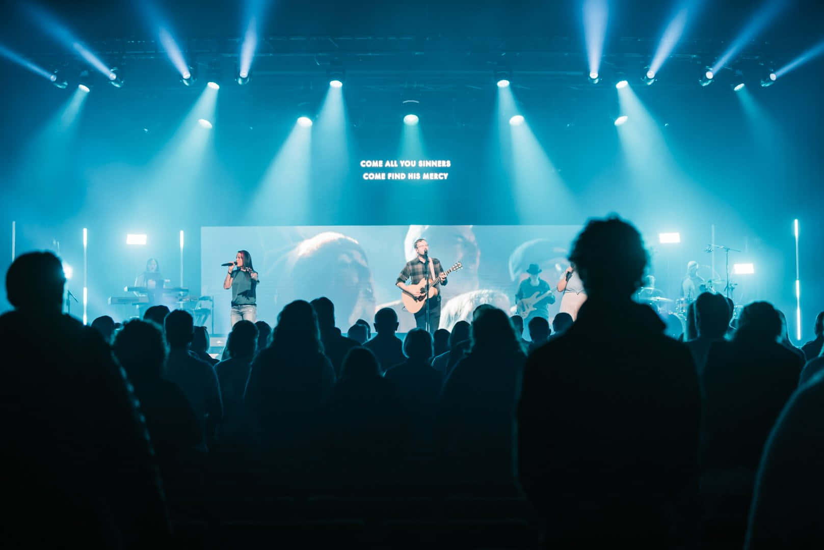 Church Stage 桌布 壁紙 — 剪影般的音樂會觀眾抬頭望向一個被冷藍色聚光燈籠罩的舞台，音樂家們在一個顯示歌詞的大螢幕下表演。