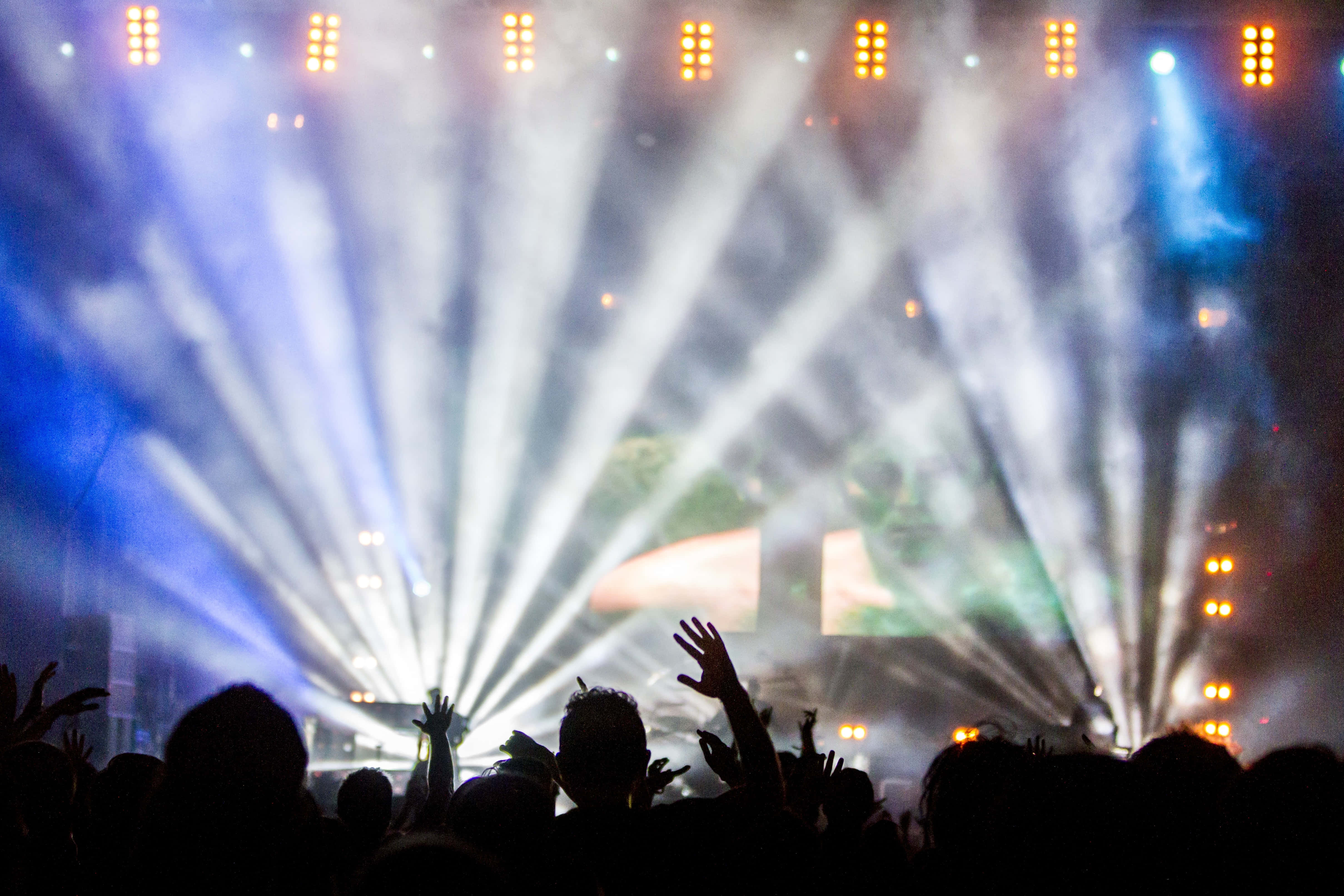 music festival 桌布 壁紙 — 剪影般的音樂會觀眾舉起雙手，朝向穿過煙霧的藍白舞台燈光射線。