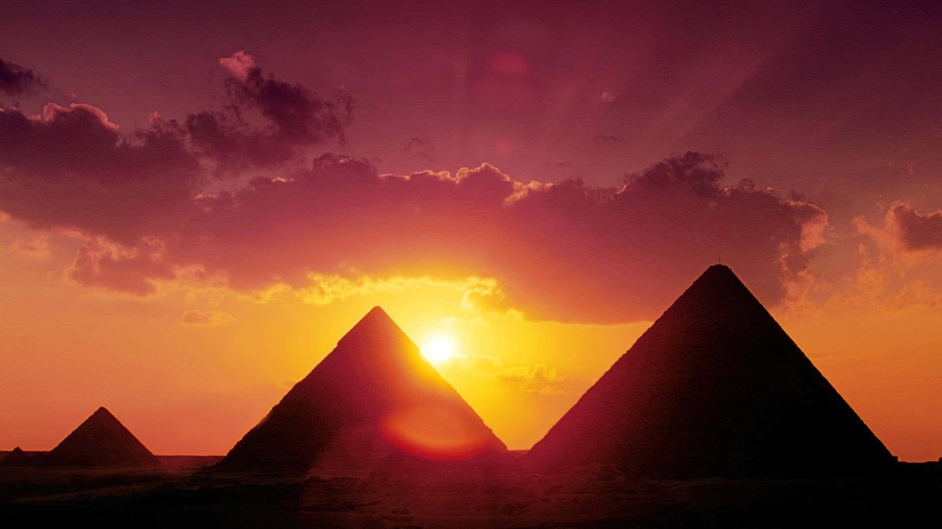 Ancient Egypt 桌布 壁紙 — 剪影金字塔在熾熱的日落天空下升起，伴隨著發光的雲彩和燦爛的太陽光芒。