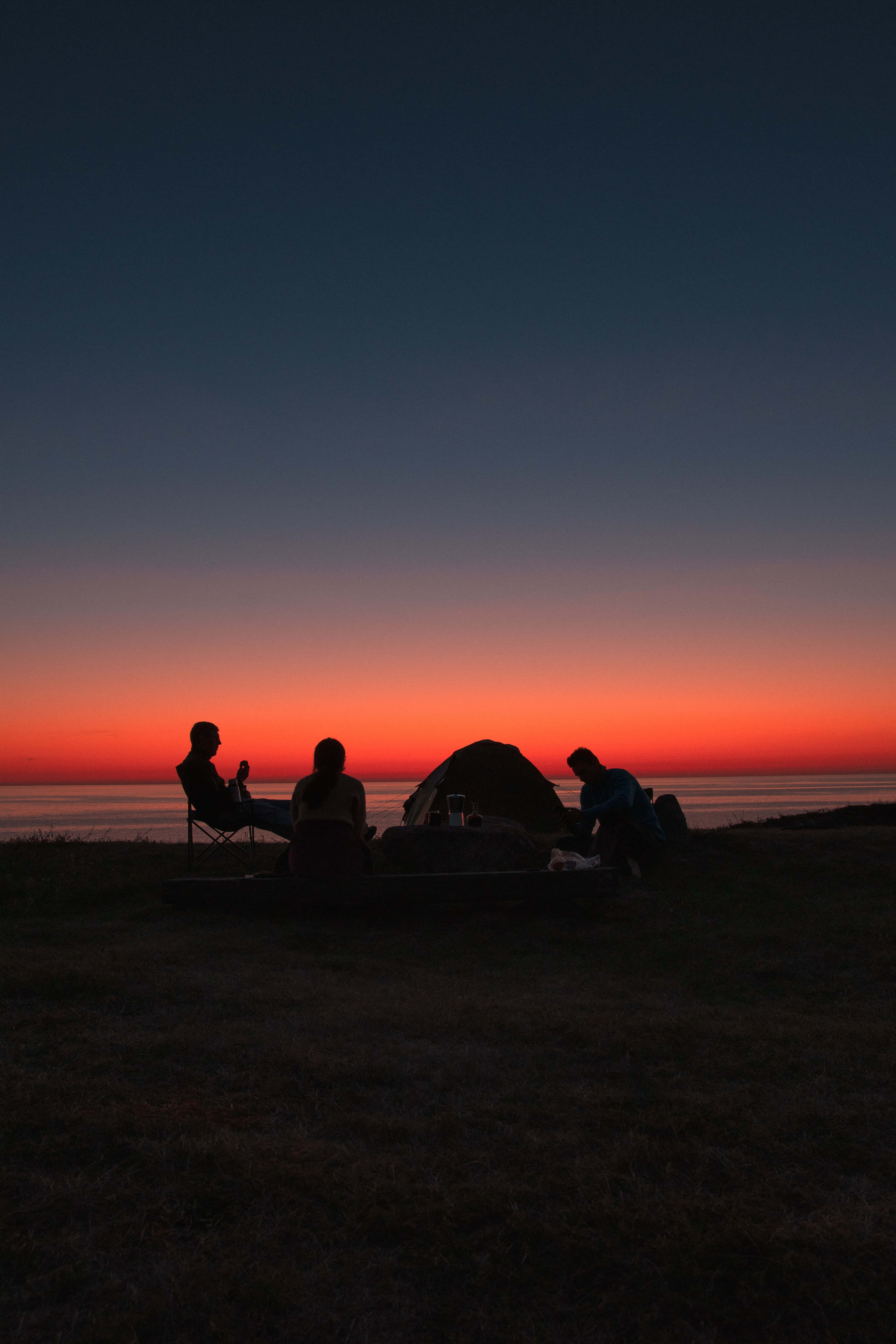Camping 桌布 壁紙 — 剪影露營者在帳篷附近放鬆，熾熱的日落將平靜水面上的地平線染上色彩。
